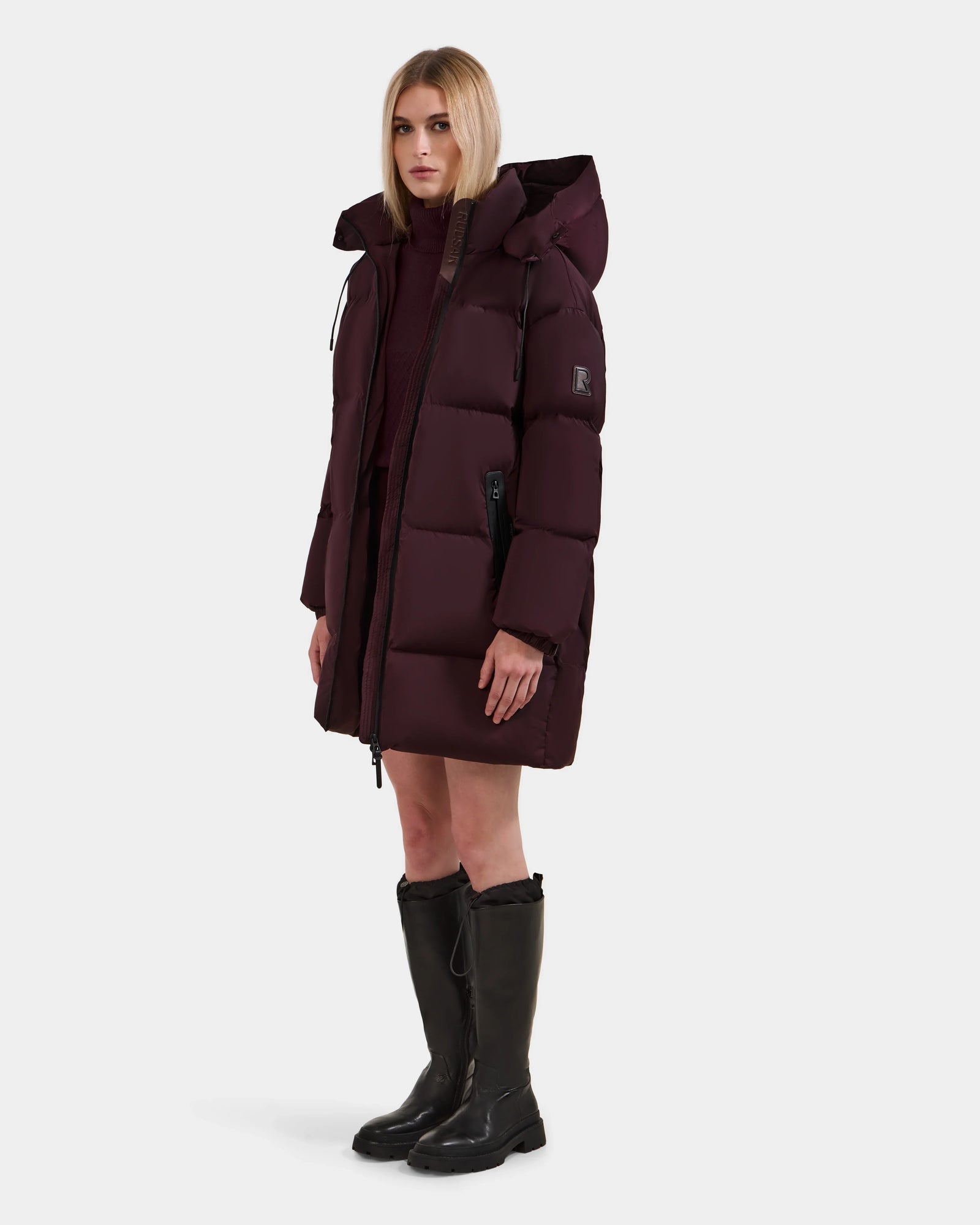 A woman faces forward against a plain white background, wearing the Rudsak KARLEE L - 8125682 Bordeaux long down puffer with hood over a dark sweater and knee-high black boots, ready for cold-weather wear.