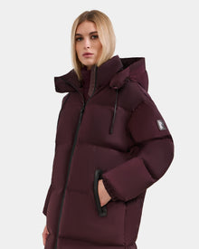 A woman wears the Rudsak KARLEE L - 8125682 Bordeaux maroon long down puffer jacket with a hood, black zipper, and sleeve patch, standing against a plain light background.