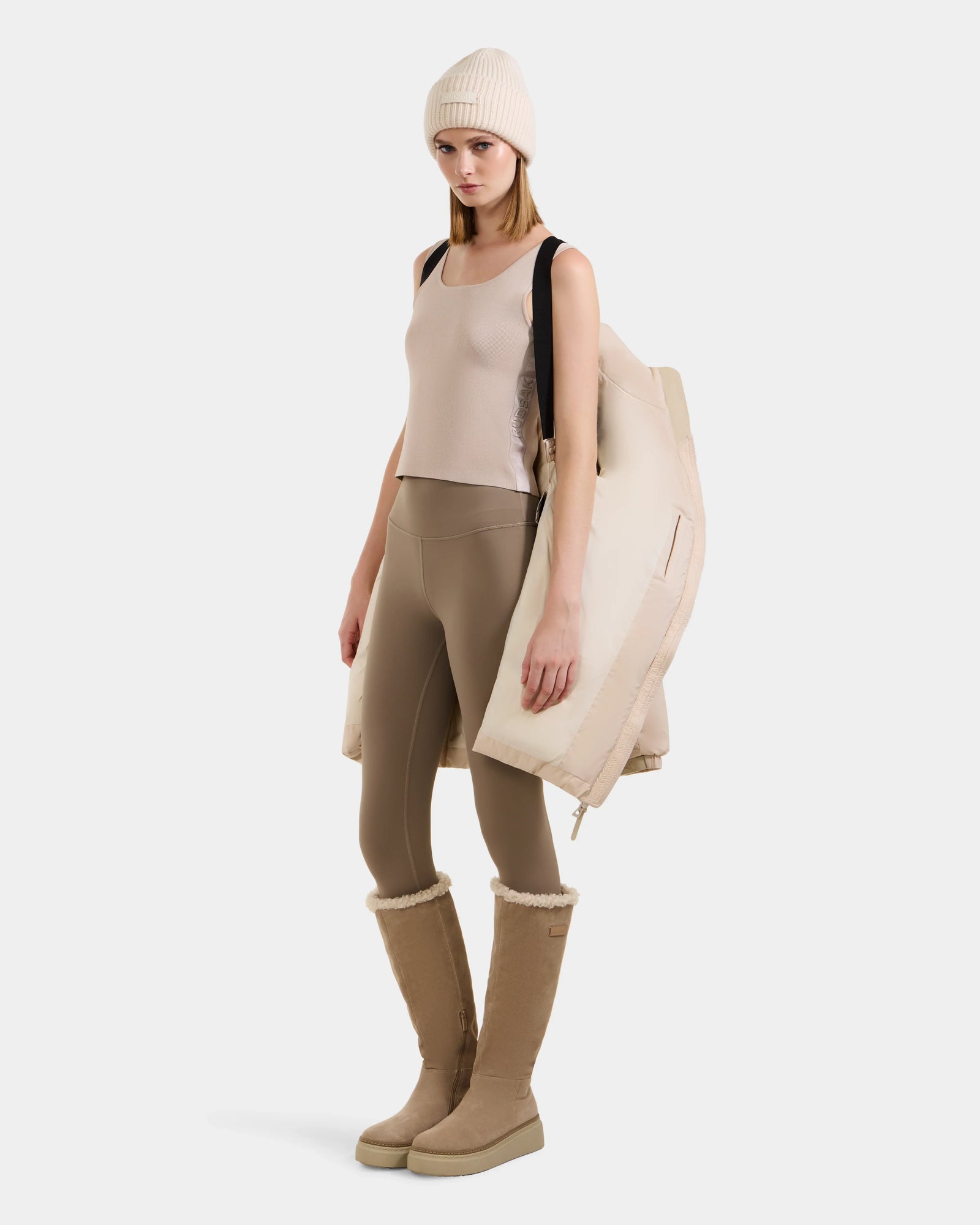 A woman stands against a white background, carrying the Rudsak KARLEE L - 8125682 Vanilla jacket with vegan leather trims over her shoulder, styled with a beige knit beanie, sleeveless top, taupe leggings, and tan knee-high boots.