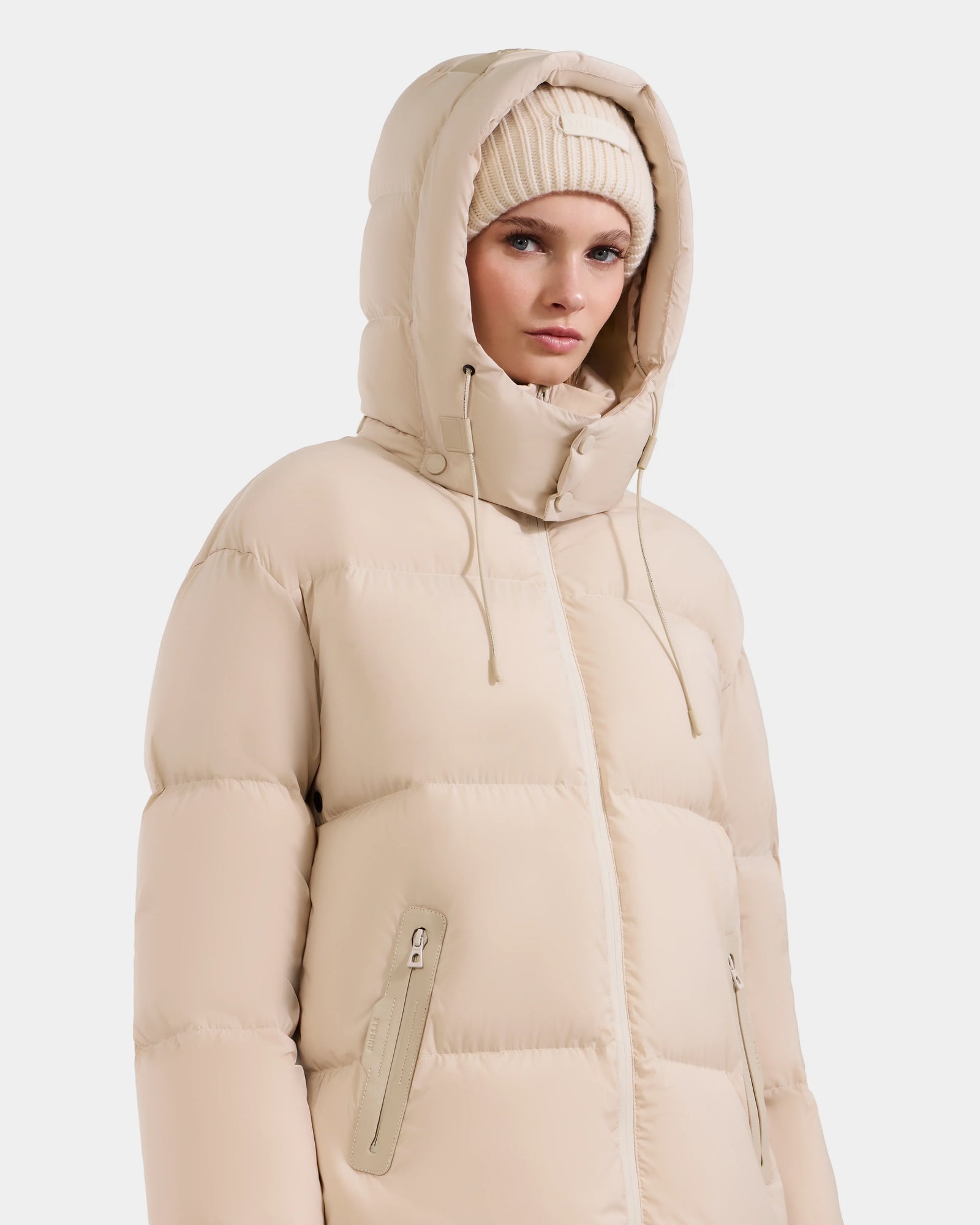 A woman wears the Rudsak KARLEE L - 8125682 Vanilla jacket, a beige puffer with vegan leather trims, drawstring hood, and zippered pockets, paired with a beige knit beanie, standing against a plain white background.