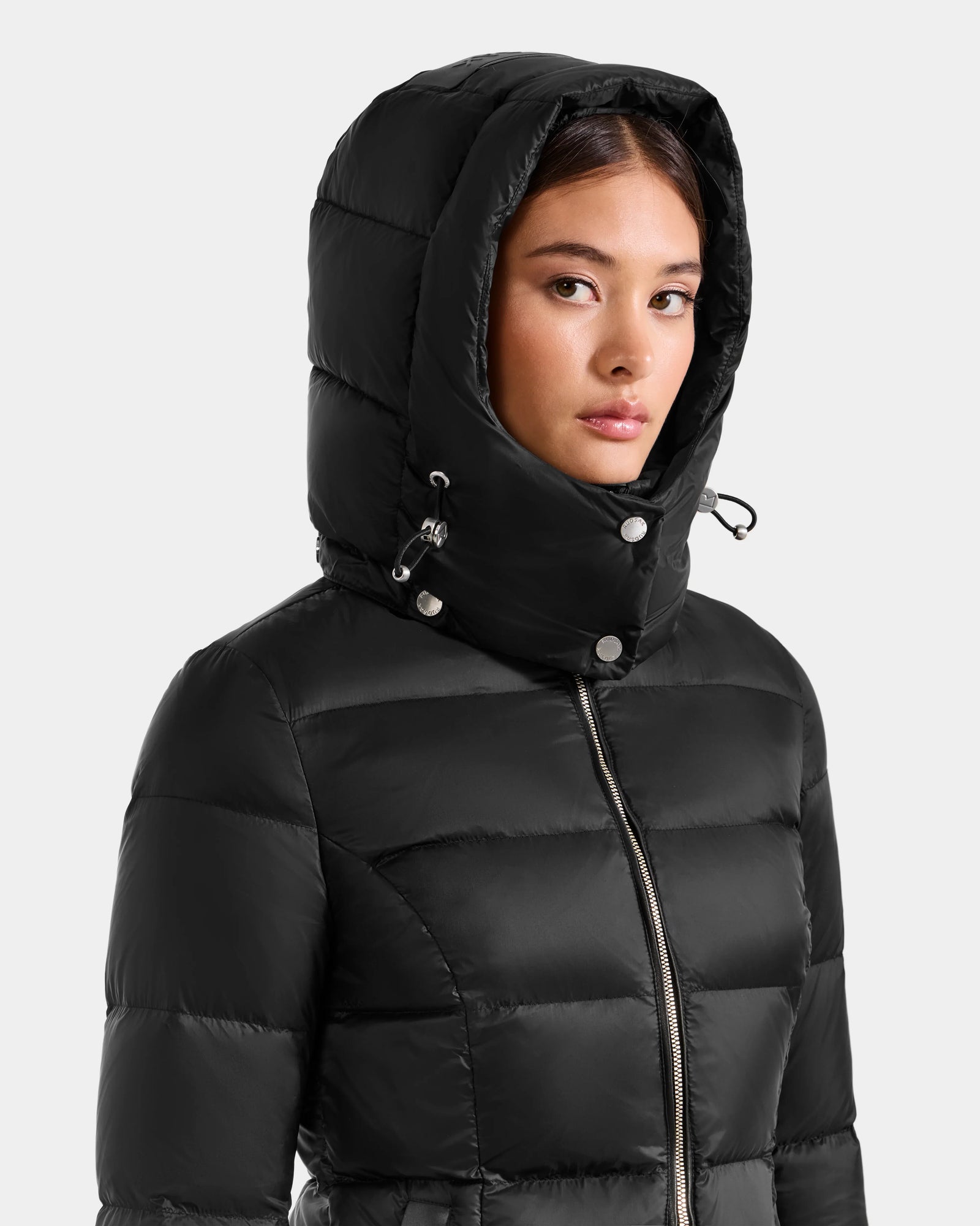 A woman in the Rudsak KATIA - L125564 Black puffer jacket with hood up, faces slightly sideways. The weather-resistant everyday jacket features a front zipper and buttoned hood. Background is light gray.