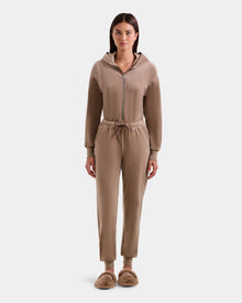 A woman stands facing forward in the Rudsak KETZY - L525503 Sand plush stretch velour zip-up hoodie and matching jogger pants with a crystal-sequin logo, plus tan fuzzy slippers, against a plain white background.