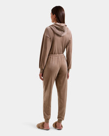 A woman with straight brown hair stands facing away against a white background, wearing the Rudsak KETZY - L525503 Sand stretch velour hooded jumper with long sleeves and jogger cuffs, paired with fluffy slippers.