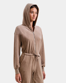 A woman wearing the Rudsak KETZY - L525503 Sand women’s velour hooded jumper, with plush stretch velour, an elastic waist, and a crystal-sequin Rudsak logo, stands against a plain light background.