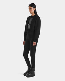A woman faces left against a white background, wearing Rudsak's LIDDIE - L525505 Black cropped crewneck sweater with a graphic design, paired with black leggings and black lace-up boots.