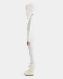 A person stands in profile wearing Rudsak's LIDDIE - L525505 Vanilla, a fitted all-white winter outfit with a fluffy white hood and tall off-white boots, set against a plain light gray background.