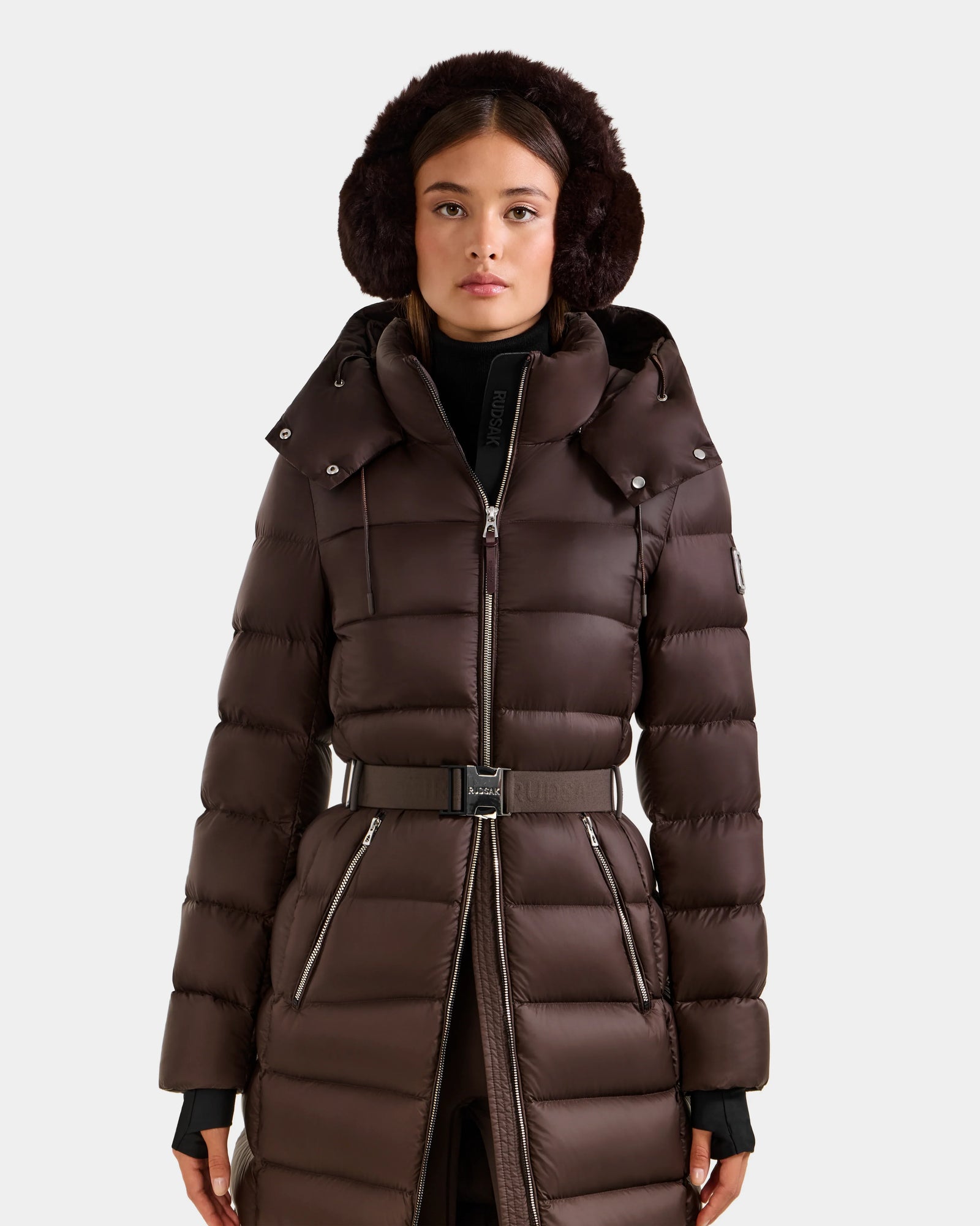 A woman stands facing forward against a light gray background, wearing the Rudsak LILITH L - L125528 Espresso dark brown maxi down puffer coat with a removable belt, black turtleneck, and dark fur earmuffs.