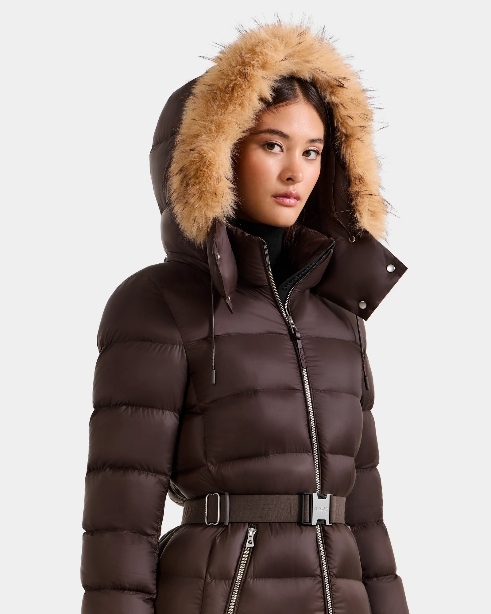 A woman wears the Rudsak LILITH L - L125528 Espresso maxi down puffer with a removable belt and light brown faux fur-trimmed hood, standing against a plain gray background.
