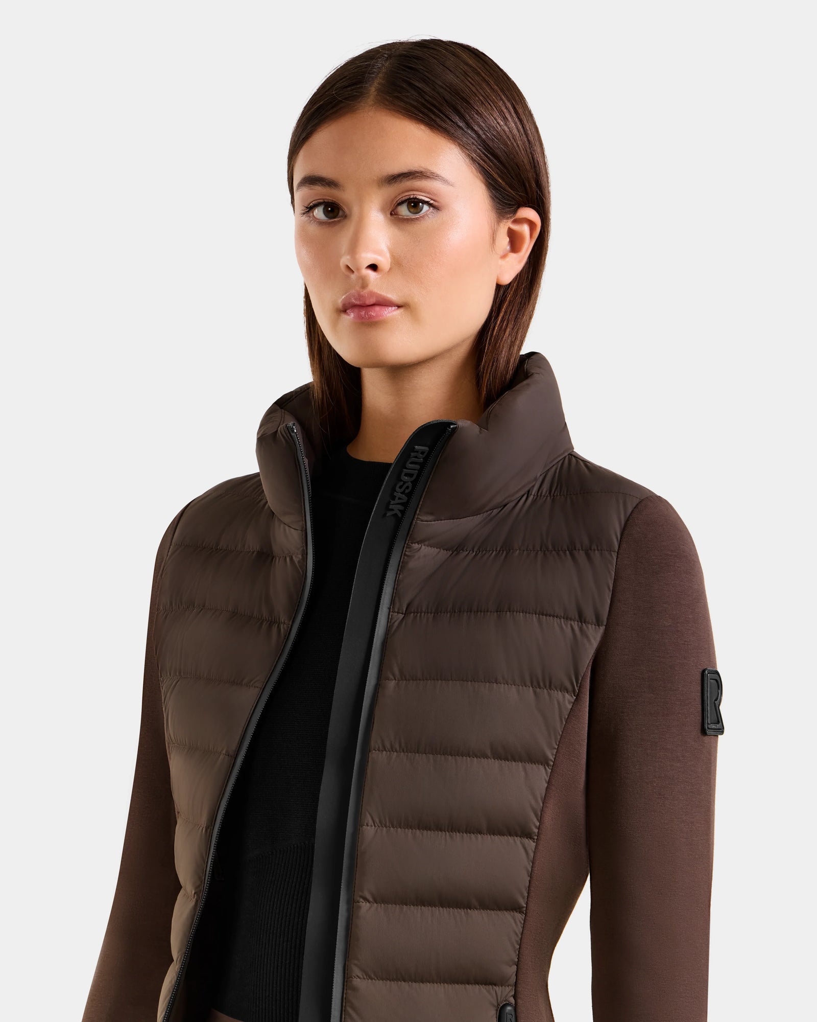A woman with straight brown hair wears the Rudsak MARRY M - L125519 Espresso women’s hybrid jacket with a high collar and down insulation over a black top, posing against a plain light background.