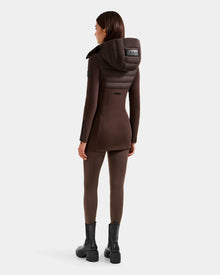 A woman faces away, wearing the Rudsak MARRY M - L125519 Espresso hybrid jacket with a large removable hood, paired with brown leggings and black ankle boots, set against a plain white background.