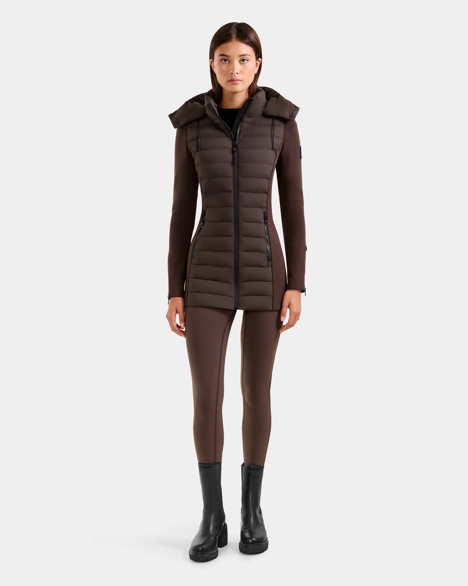 A woman stands facing forward, wearing the Rudsak MARRY M - L125519 Espresso women’s hybrid jacket with down insulation and a removable hood, brown leggings, and black ankle boots against a plain light background.