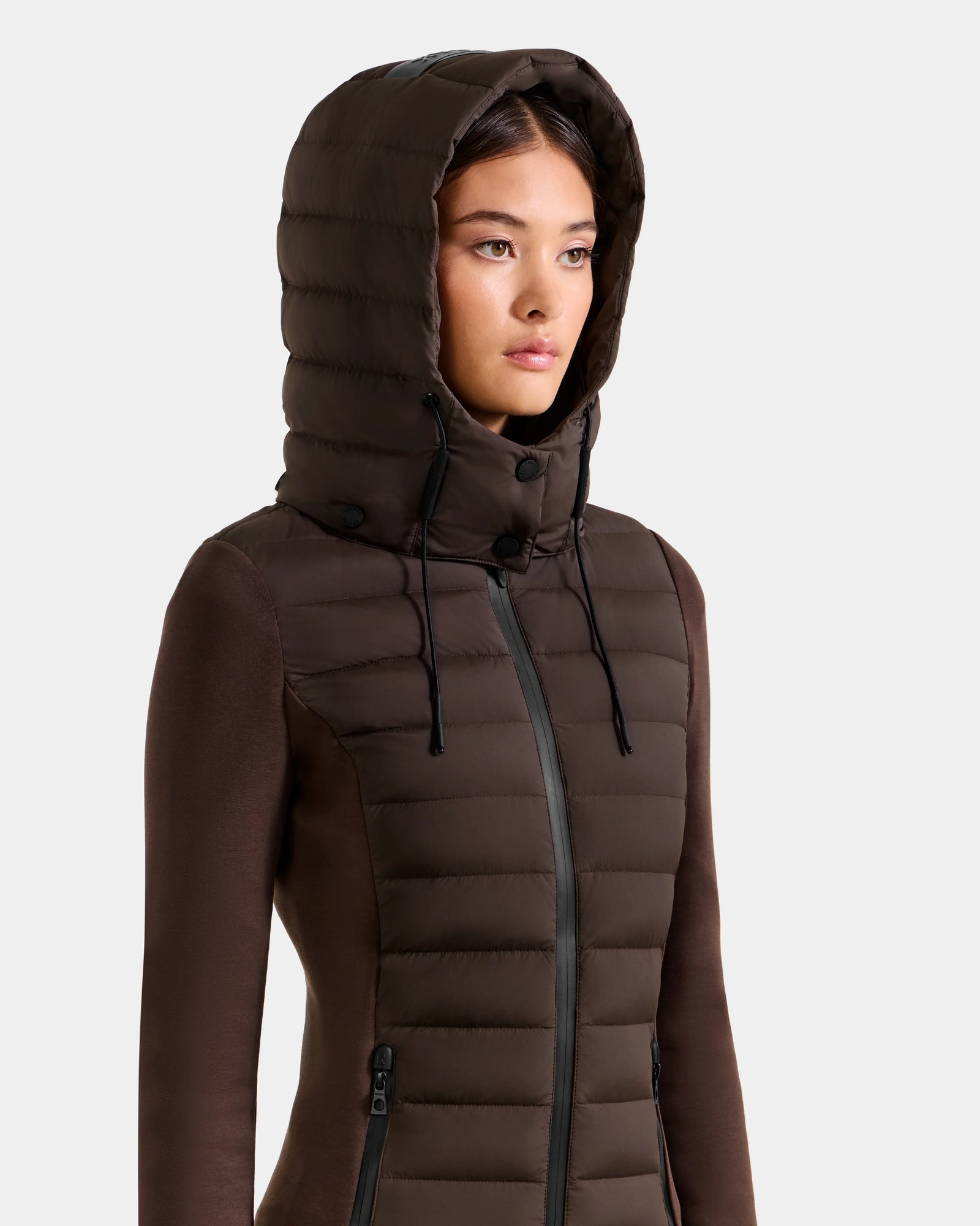 A woman wears the Rudsak MARRY M - L125519 Espresso hybrid jacket, featuring a fitted brown design with a large padded removable hood, down insulation, front zipper, and zippered side pockets against a plain light background.