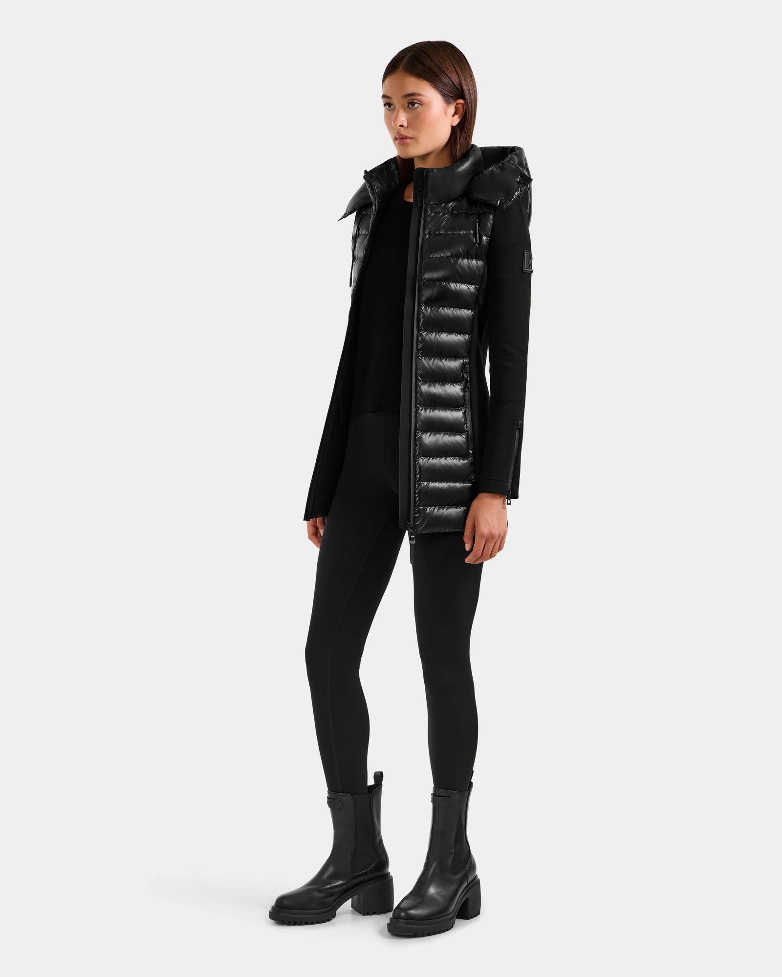 A woman wears the Rudsak MARRY S - L125521 Black long weather-resistant puffer coat with a high collar, paired with black leggings, boots, and top against a plain light gray background.