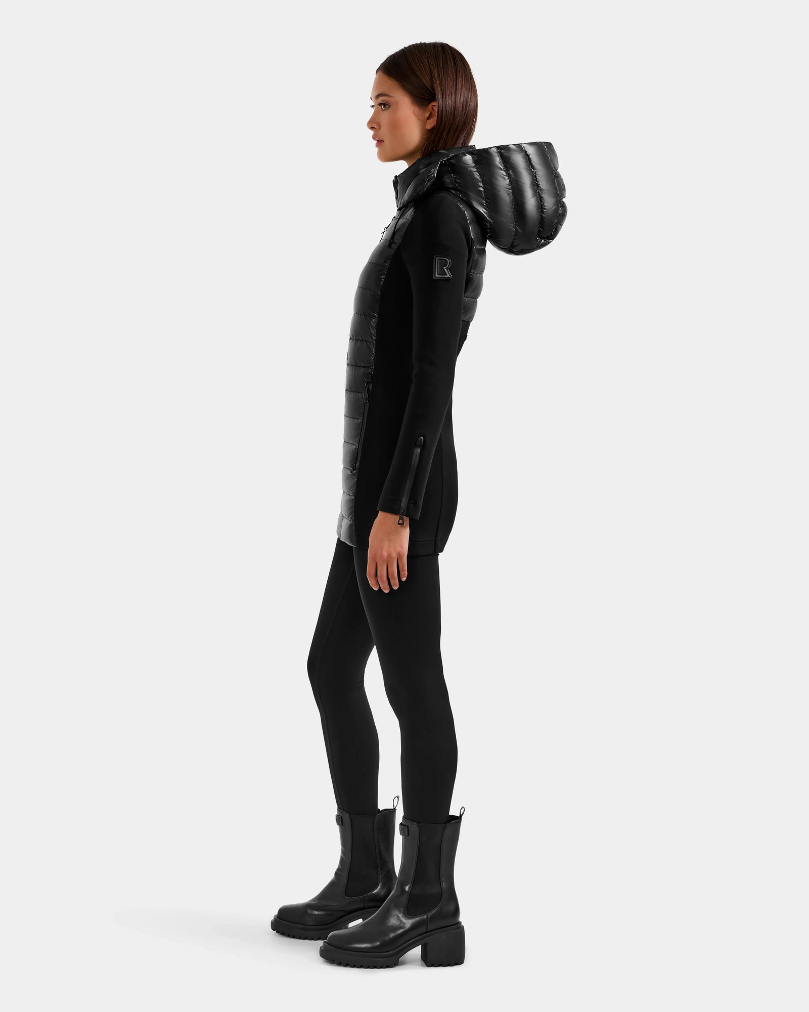 A woman stands in profile against a light gray background, wearing the Rudsak MARRY S - L125521 Black weather-resistant puffer coat with RDS-certified down, paired with black leggings and heeled boots.
