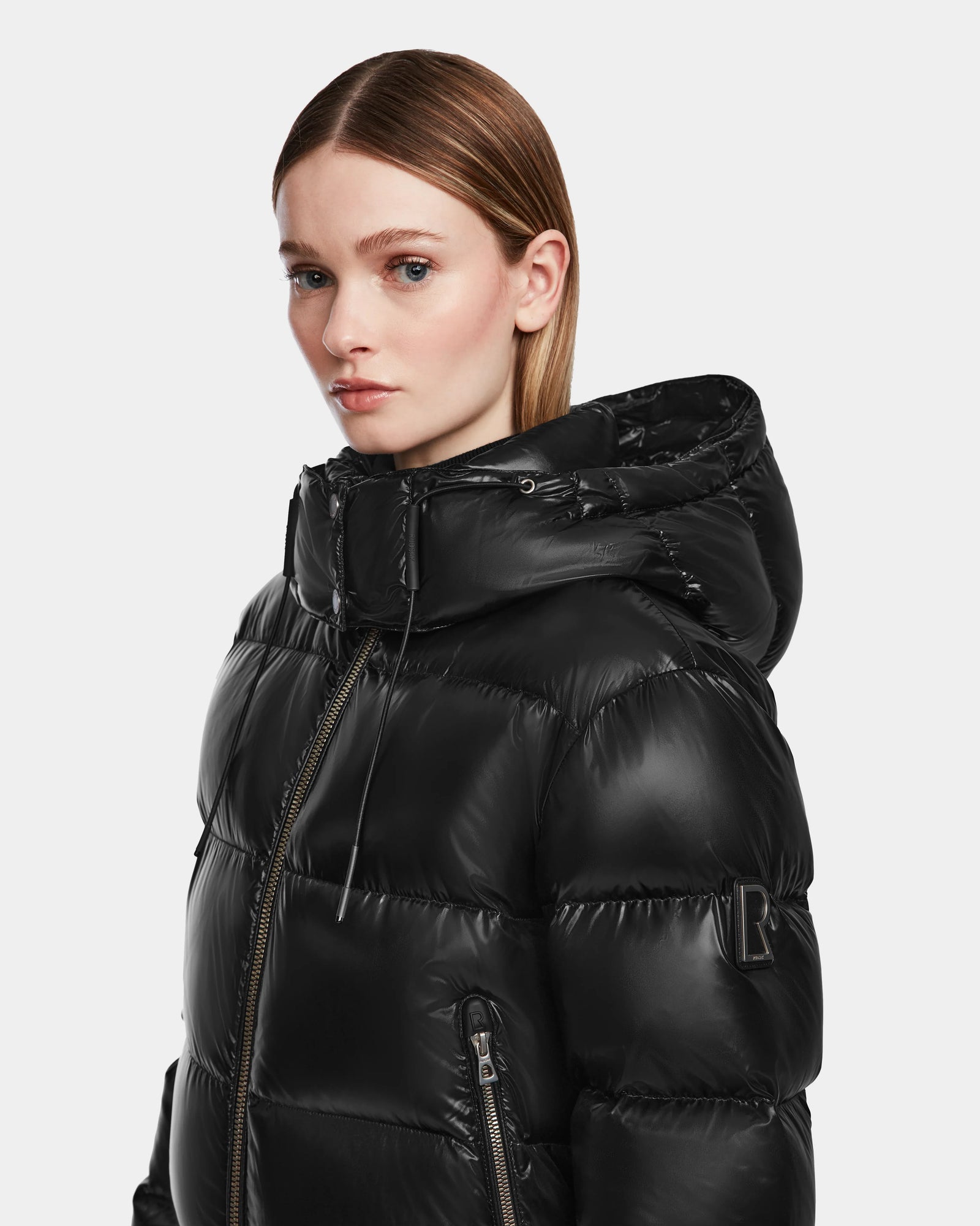 A young woman with straight, light brown hair models the Rudsak MAUDE Q-S - L124553 Black, a shiny winter jacket with a hood. She faces slightly to the side against a plain light background.