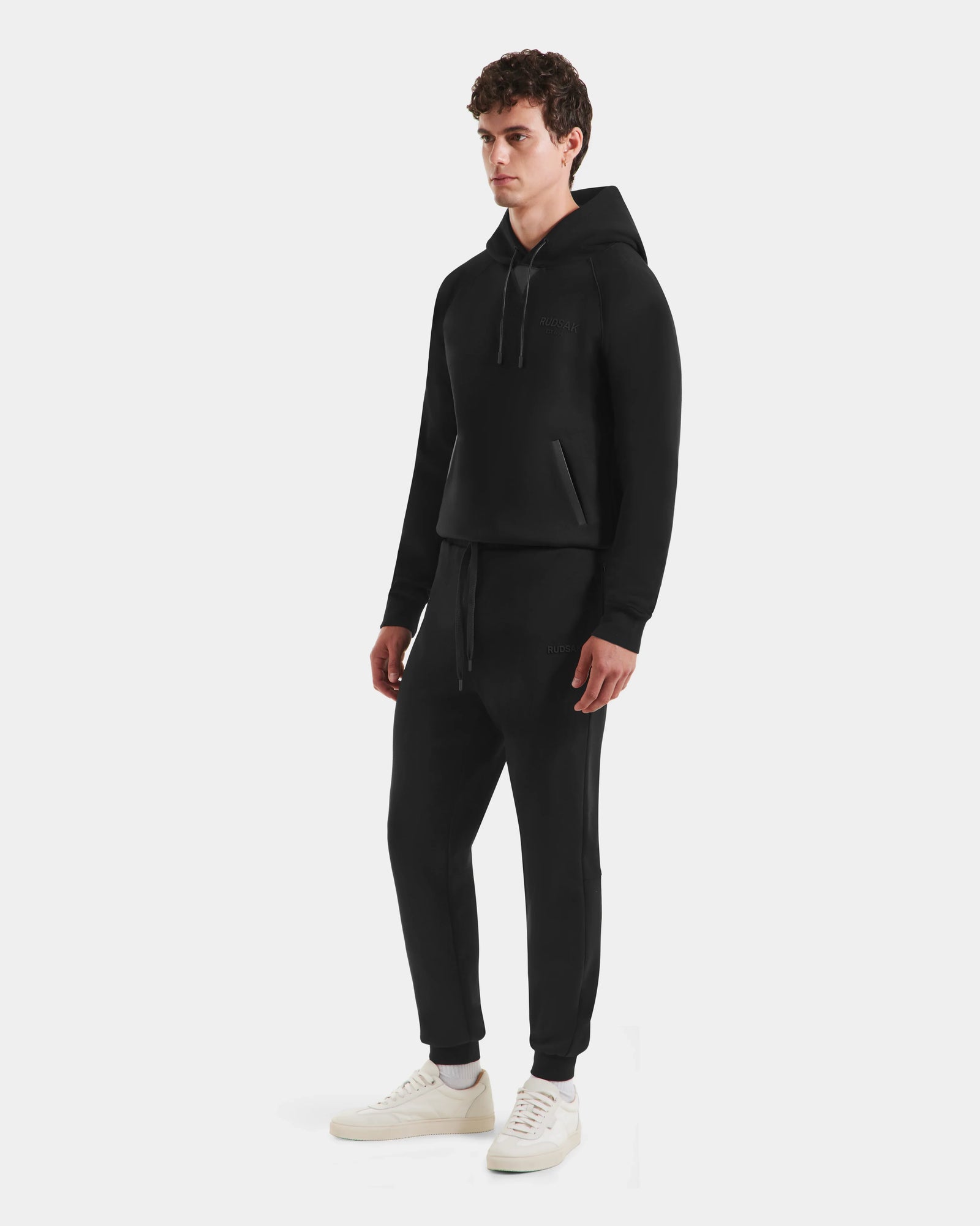 A man stands against a white background wearing the Rudsak MAXTON - U525007 Black M hoodie, paired with unisex cotton-blend joggers and white sneakers. He looks slightly to the side, hands relaxed by his sides.