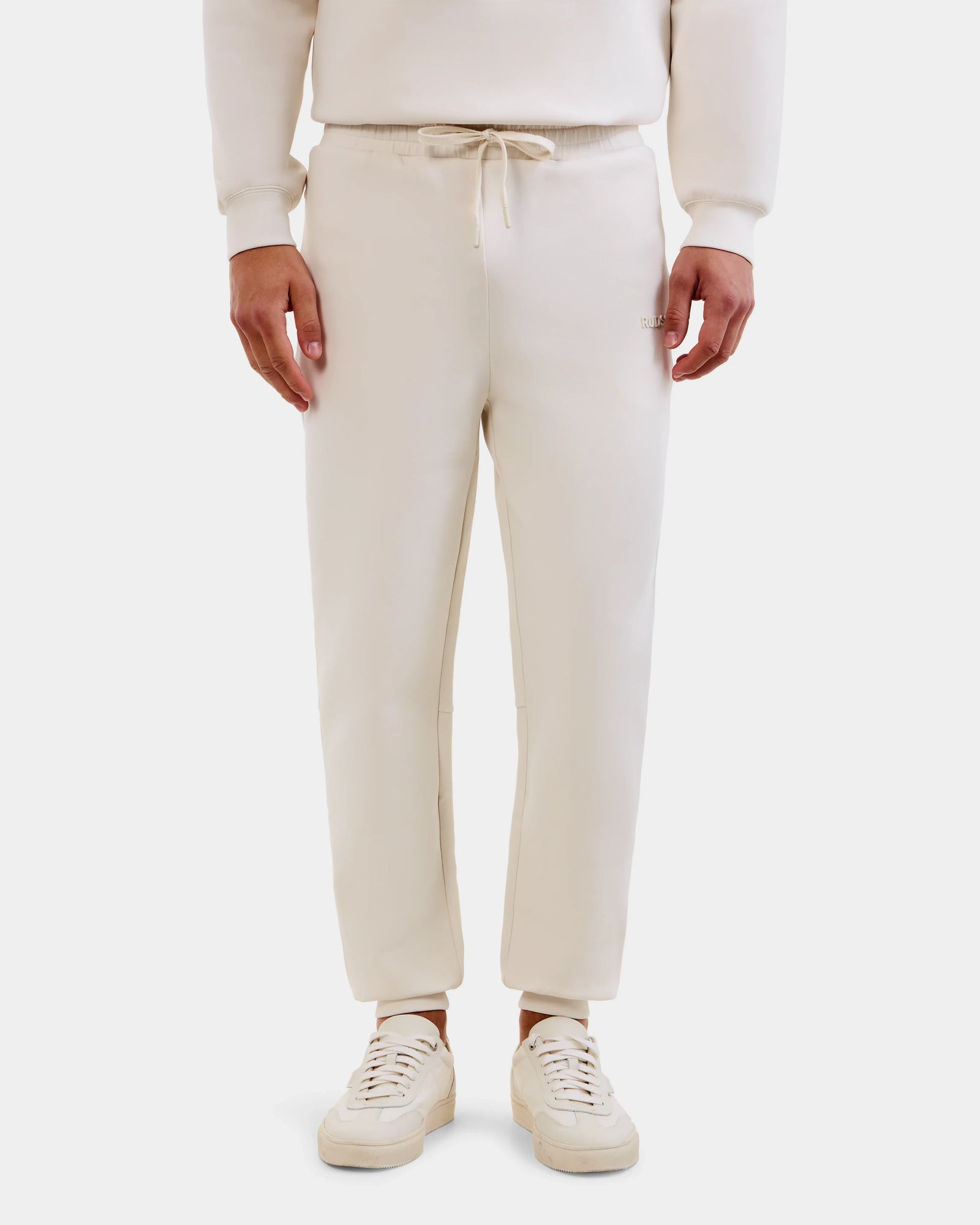 A person wears the Rudsak MAXTON - U525007 Vanilla M jogger pants, shown from the waist down against a white background, highlighting everyday comfort in this cream cotton-blend unisex jogger style.