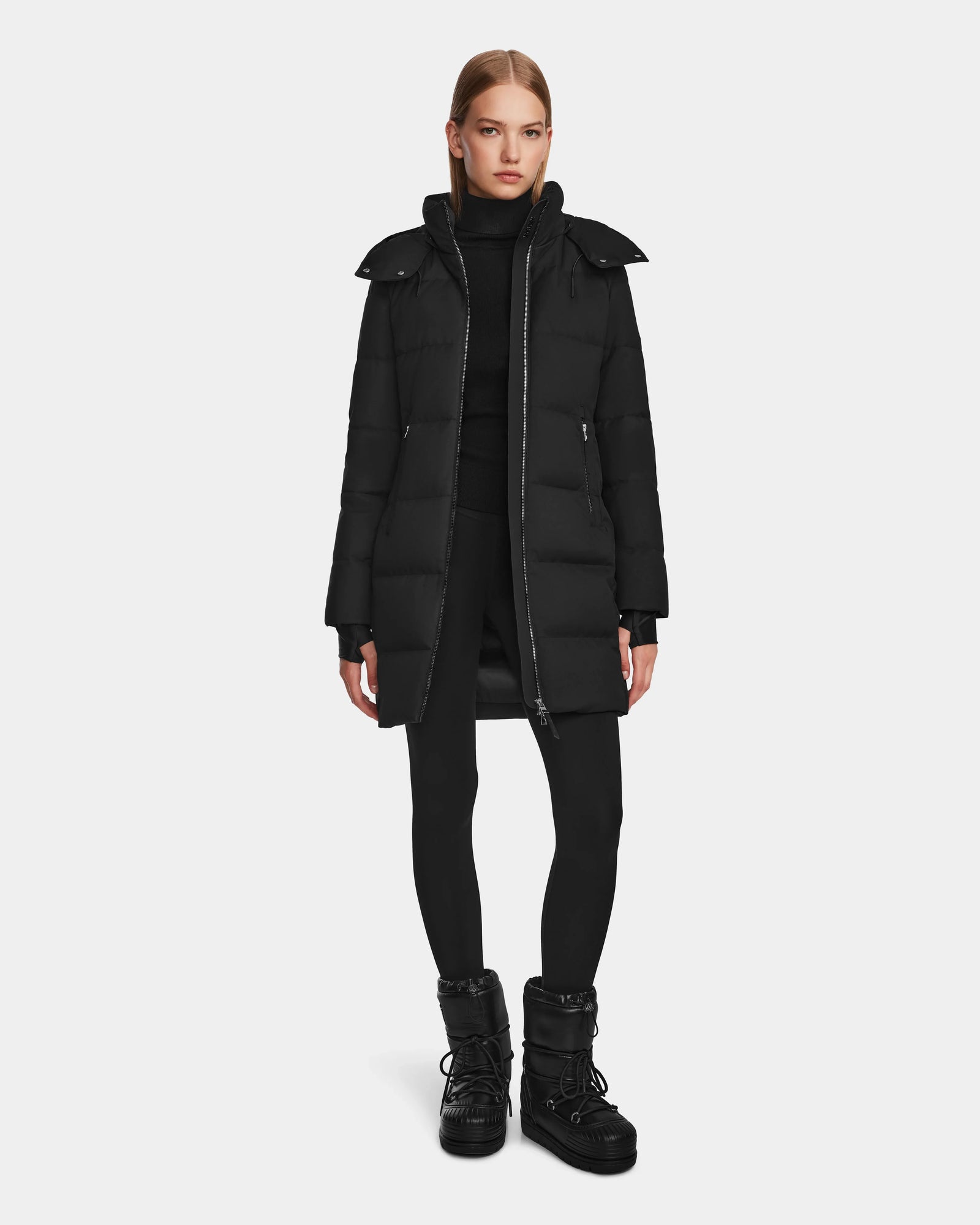 A woman stands facing forward in the Rudsak MIRIAM - L123544 Black womens down puffer coat with RDS-certified down, paired with a black turtleneck, leggings, and winter boots, set against a plain white background.