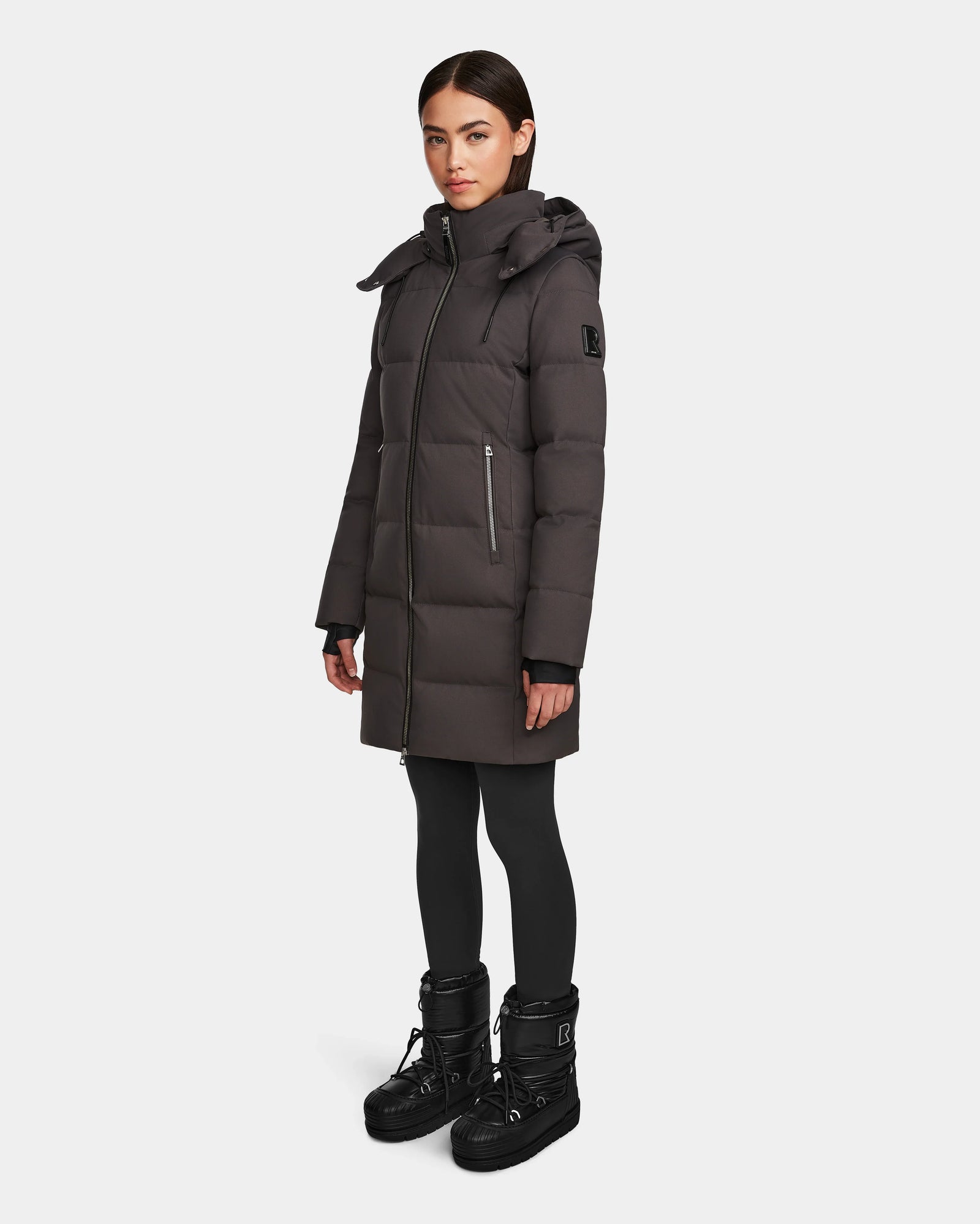 A woman wearing the Rudsak MIRIAM - L123544 Grey down puffer coat with a hood, paired with black leggings and winter boots, stands against a white background—ideal for refreshing your winter wardrobe.