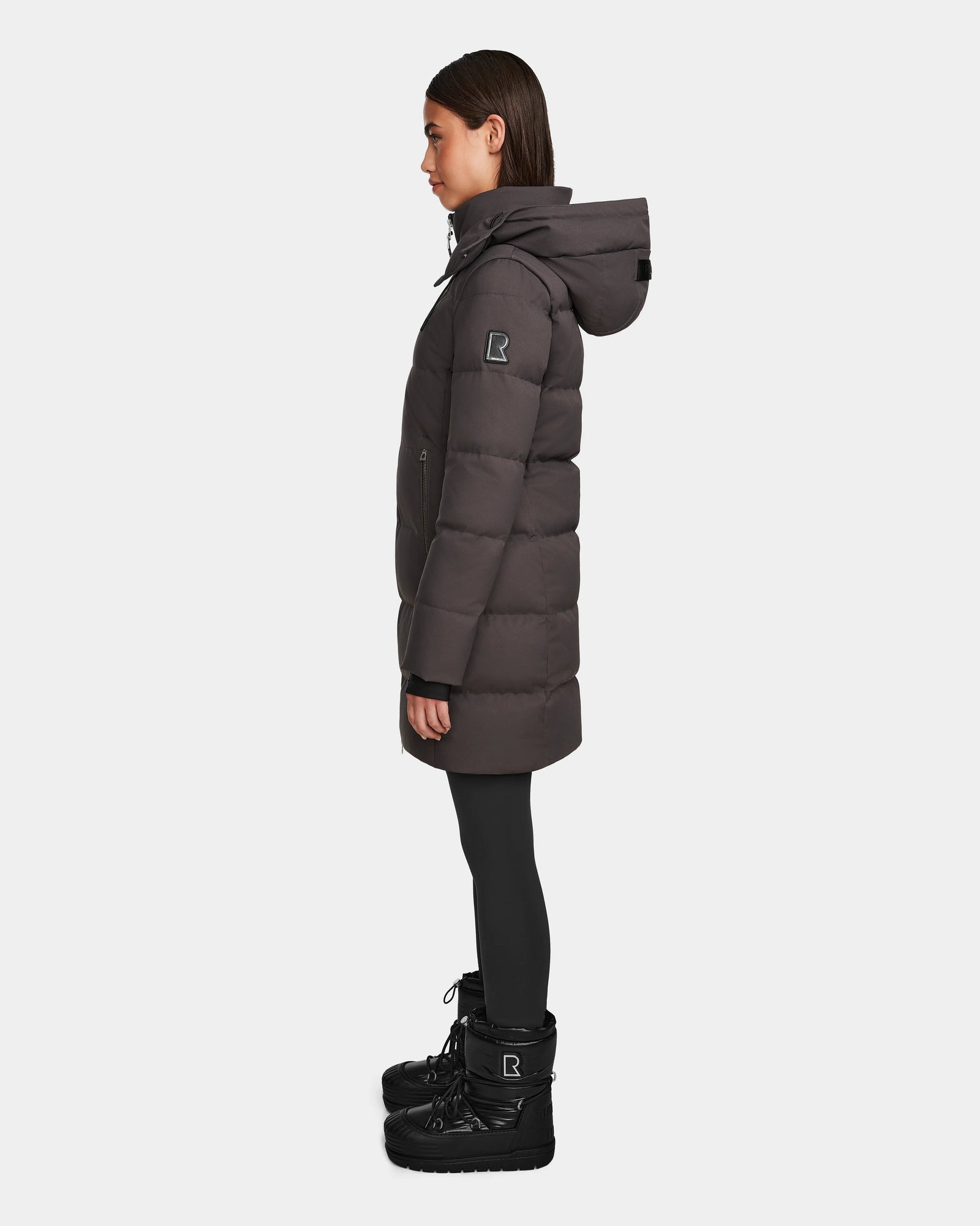 A woman in profile showcases the Rudsak MIRIAM - L123544 Grey down puffer coat with a hood, paired with black leggings and winter boots—highlighting a sleek winter look against a plain white background.