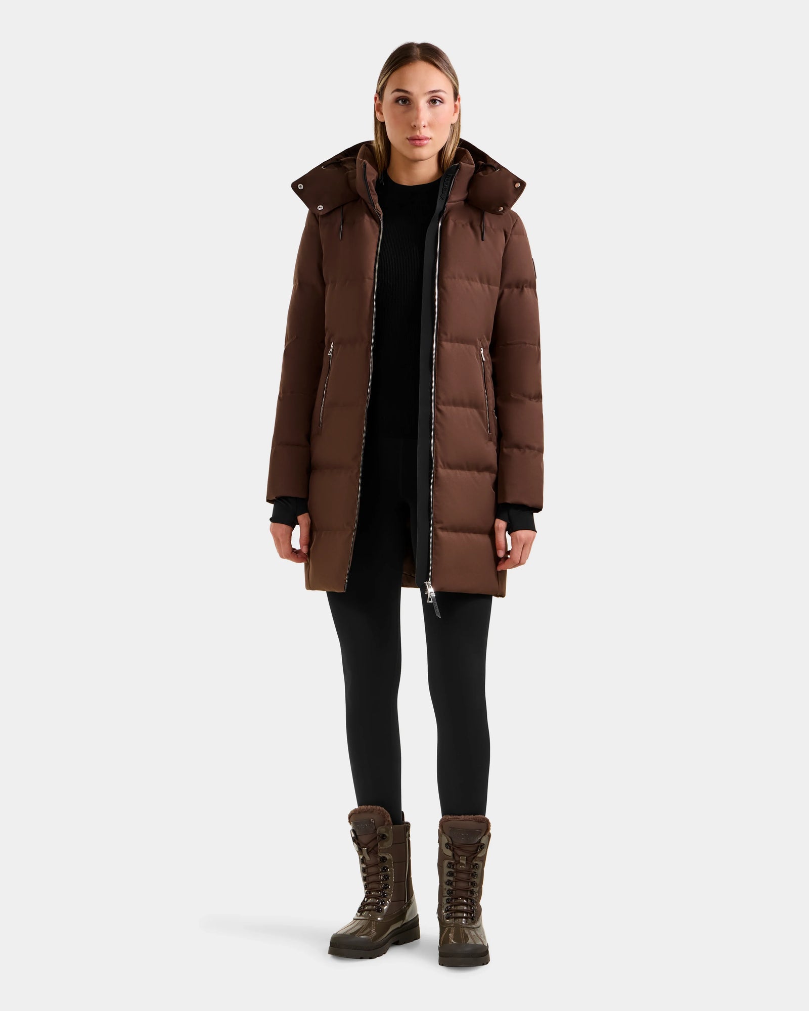 A woman faces forward in the Rudsak MIRIAM - L123544 Mocha down puffer coat with a weather-resistant shell, worn unzipped to show a black top. She pairs it with black leggings and brown lace-up winter boots against a light gray background.