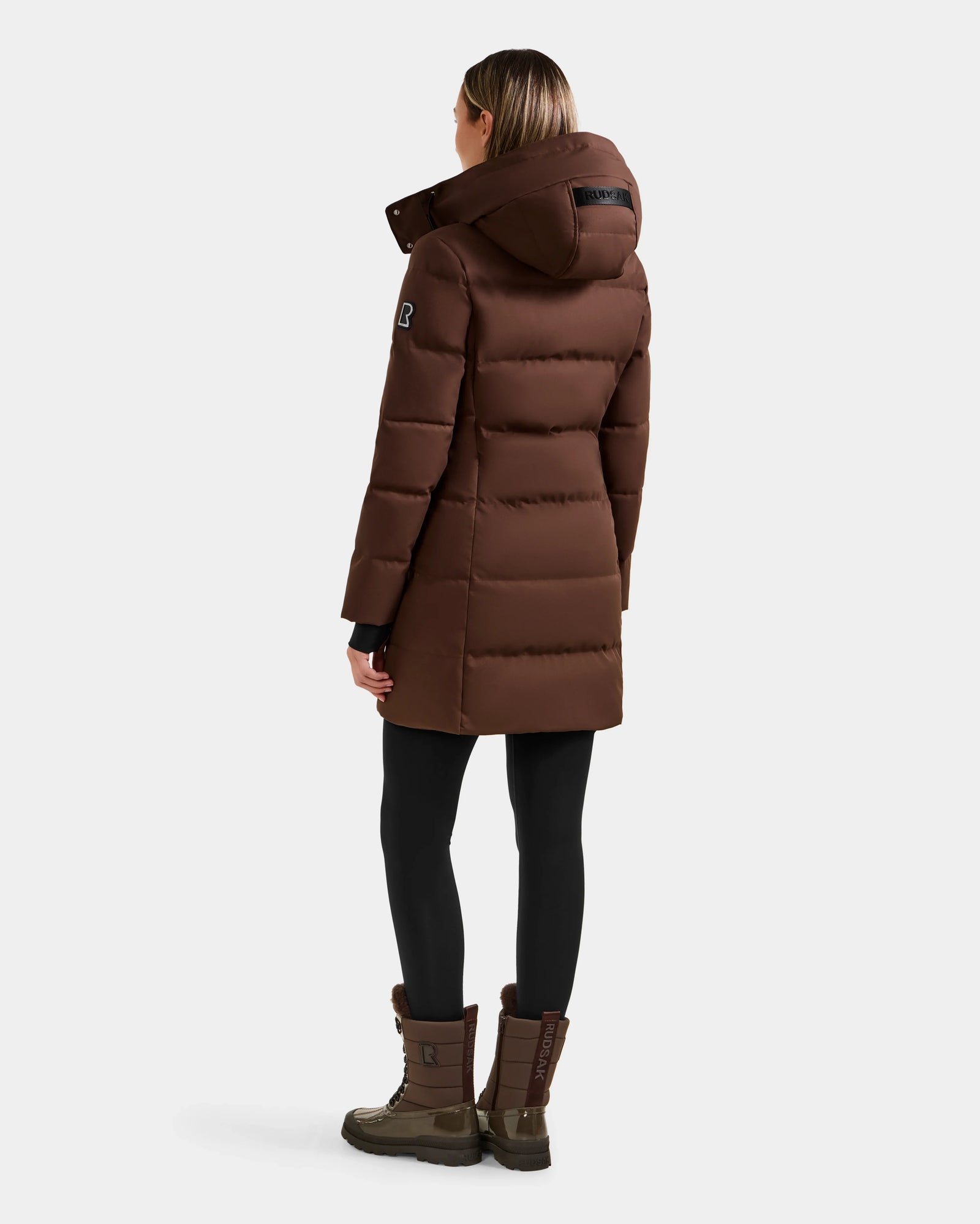 A person models the Rudsak MIRIAM - L123544 Mocha women’s down puffer coat with a hood, paired with black leggings and brown boots, standing against a plain light background to showcase the back and side of the outfit.
