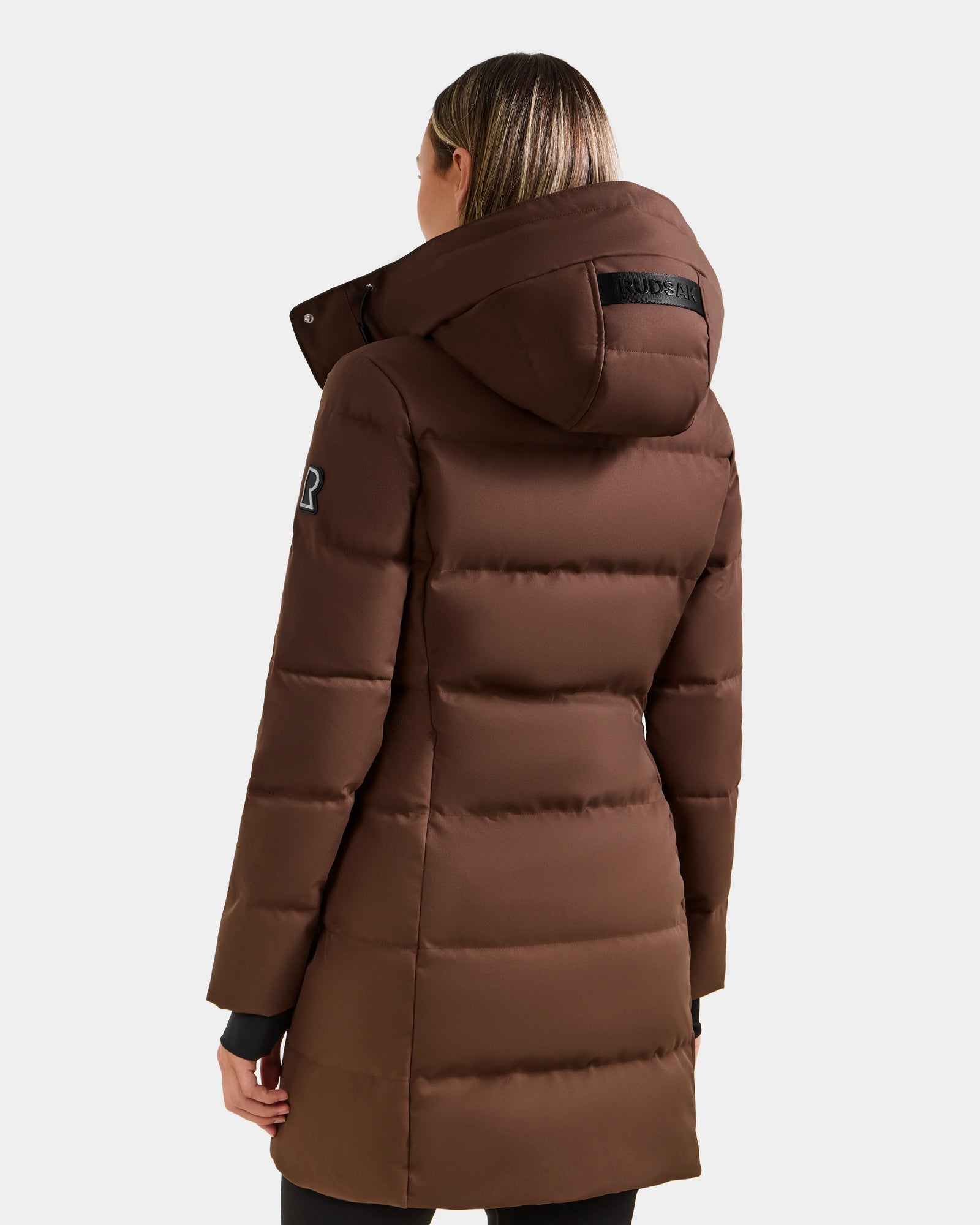 A person is seen from the back wearing the Rudsak MIRIAM - L123544 Mocha women's down puffer coat, featuring a hood, horizontal quilting, an upper-arm patch, and a weather-resistant shell against a plain light background.