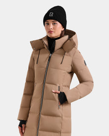 A woman stands facing forward in the Rudsak MIRIAM - L123544 Sand puffer coat, featuring a hood and zipper pockets. She wears a black knit beanie with a white R logo and a black turtleneck. The background is plain white.