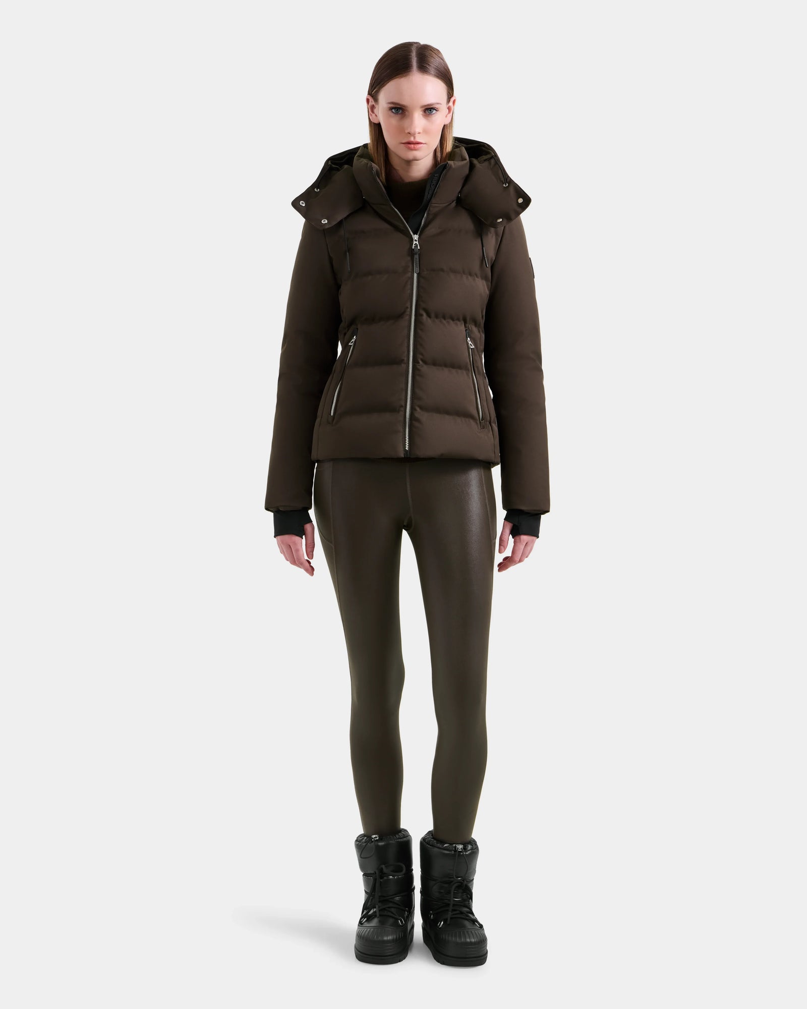 A woman faces forward, wearing the Rudsak MIRRY M - L124533 Espresso RDS-certified down puffer jacket with a hood, olive green leggings, and black platform boots against a plain white background.