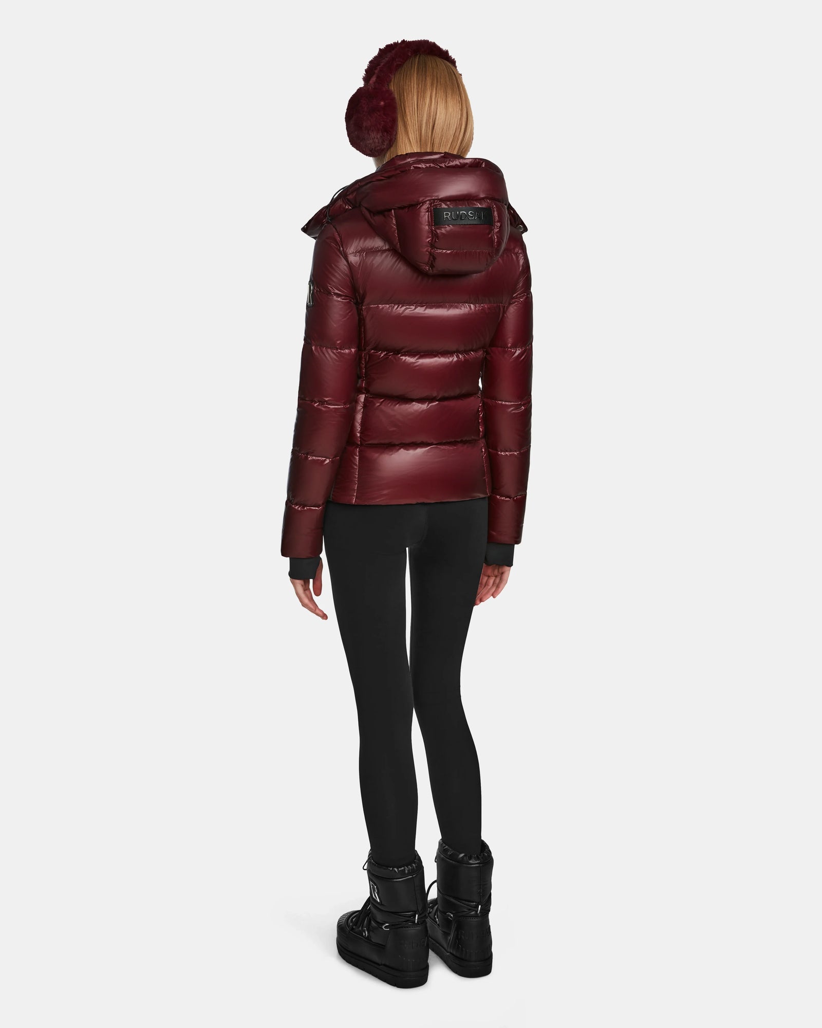 A woman in the Rudsak MIRRY S - L123545 Wine puffer jacket, black leggings, black winter boots, and maroon earmuffs stands with her back to the camera against a plain white background.