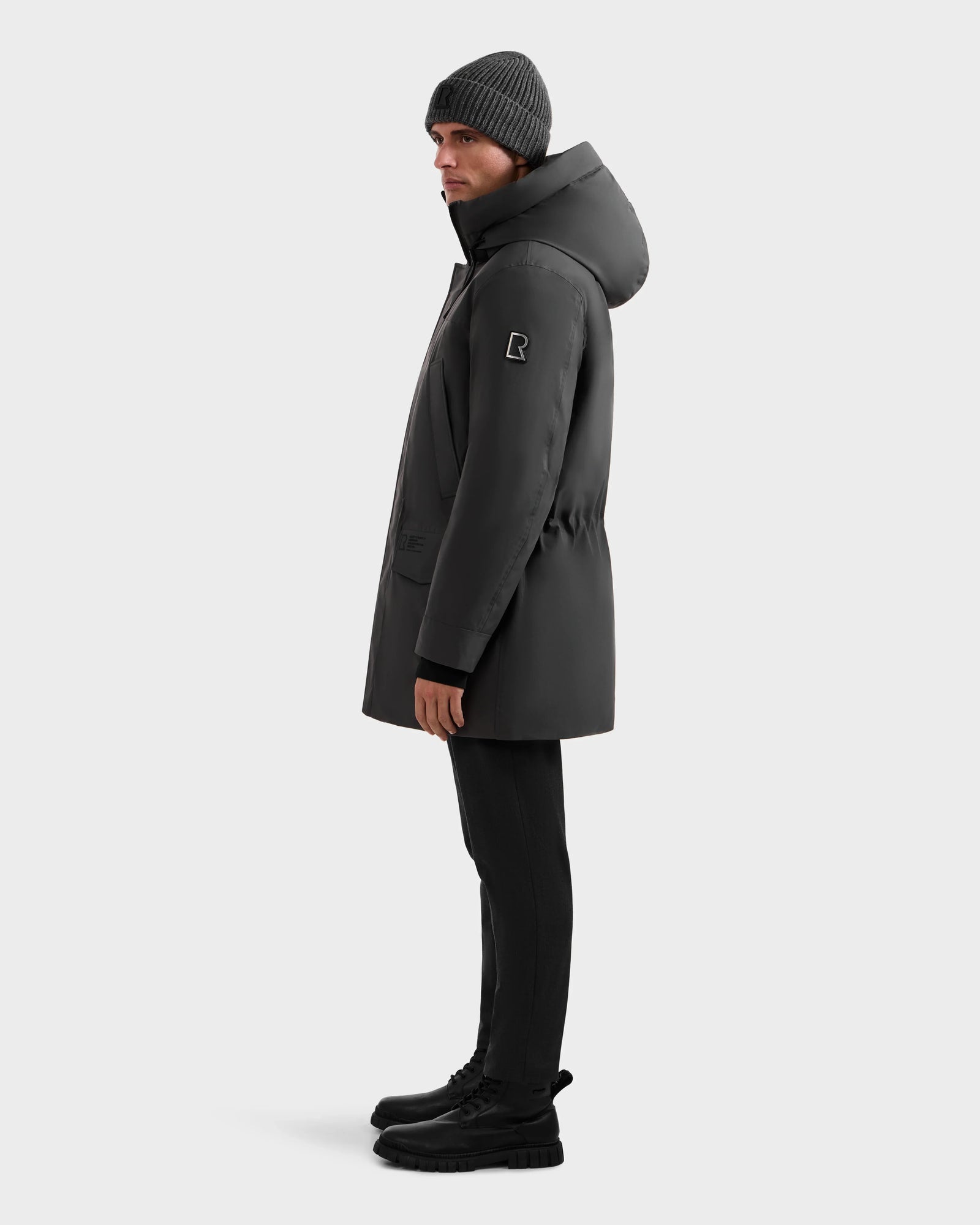 A person in a dark winter coat, black pants, black boots, and a gray knit beanie stands in profile against a light background—showcasing the Rudsak MITCHELL - M125513 Grey men’s down parka, a true winter essential.