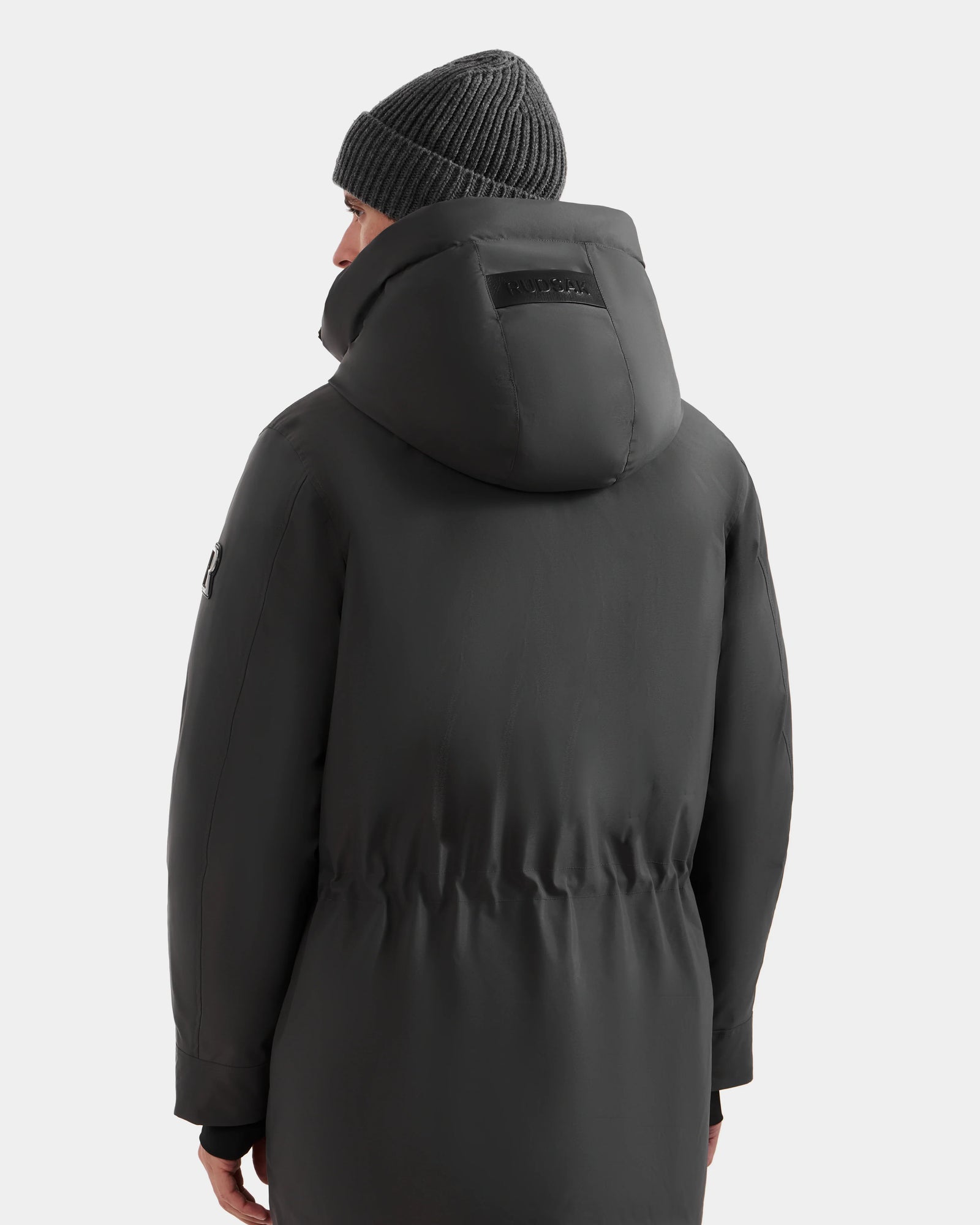 A person in the Rudsak MITCHELL - M125513 Grey men’s down parka, featuring RDS-certified down, and a ribbed knit beanie faces away from the camera, highlighting the back of this winter essential against a plain light background.