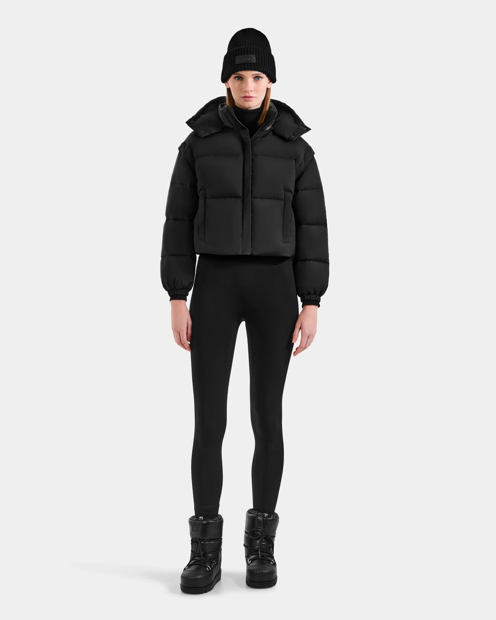 A woman stands facing forward wearing the Rudsak OPRAH - L125546 Black women’s cropped down puffer with RDS-certified down, styled with black leggings, boots, and a knit beanie against a plain light background.