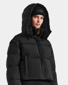 A woman in the Rudsak OPRAH - L125546 Black women’s cropped down puffer with a hood and a black knit beanie, gazing slightly to the side against a plain light gray background.