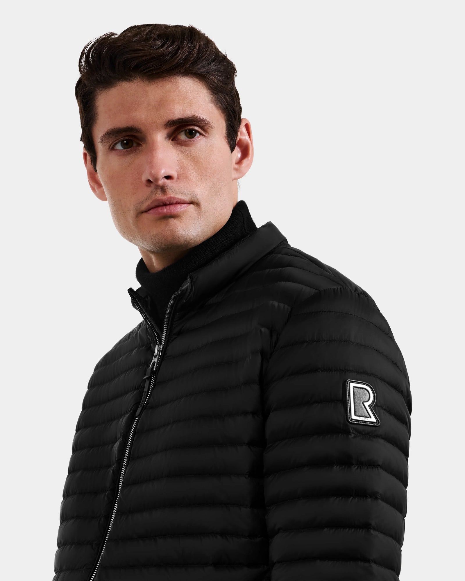 A man with short brown hair wears the Rudsak REYMOND M - M125523 Black down jacket, crafted from recycled nylon with an R logo patch on the sleeve, looking slightly left against a plain light background.