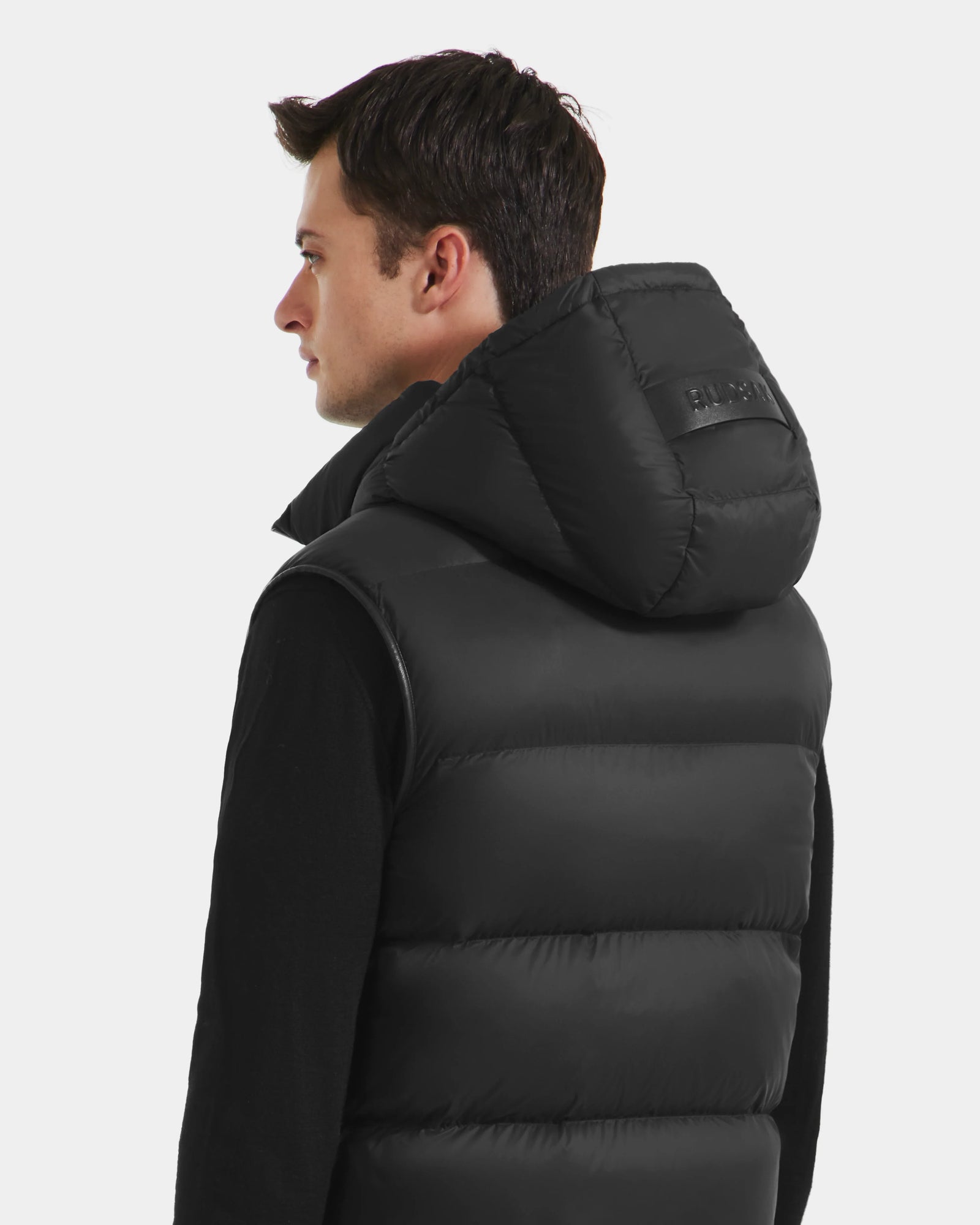 A man, seen from behind and slightly to the side against a plain white background, wears the Rudsak RICHARDSON - M125527 Black men’s hooded vest with lightweight warmth and a water-repellent finish.