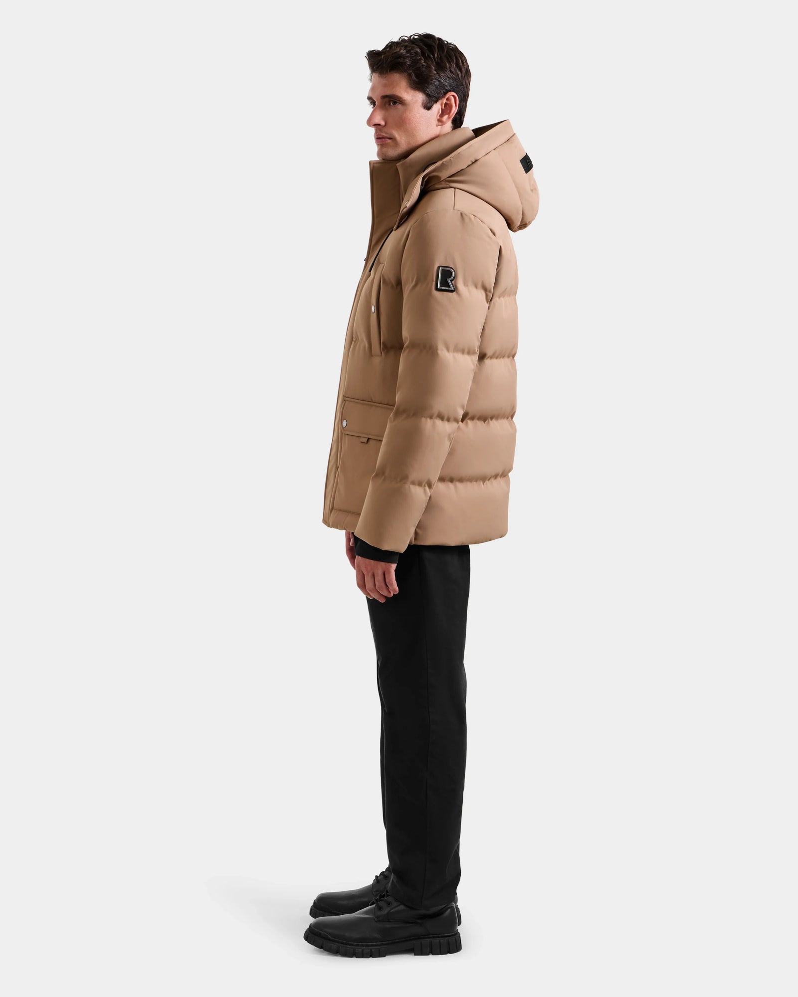A man in profile wears the RICKY - M123502 Sand men's quilted down parka by Rudsak with a hood, black pants, and black shoes against a plain white background—ideal for winter fashion.