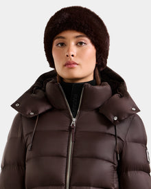 A woman in the Rudsak SANDIE - L425552 Espresso dark brown puffer jacket with a hood and matching faux fur headband gazes ahead against a plain light background.