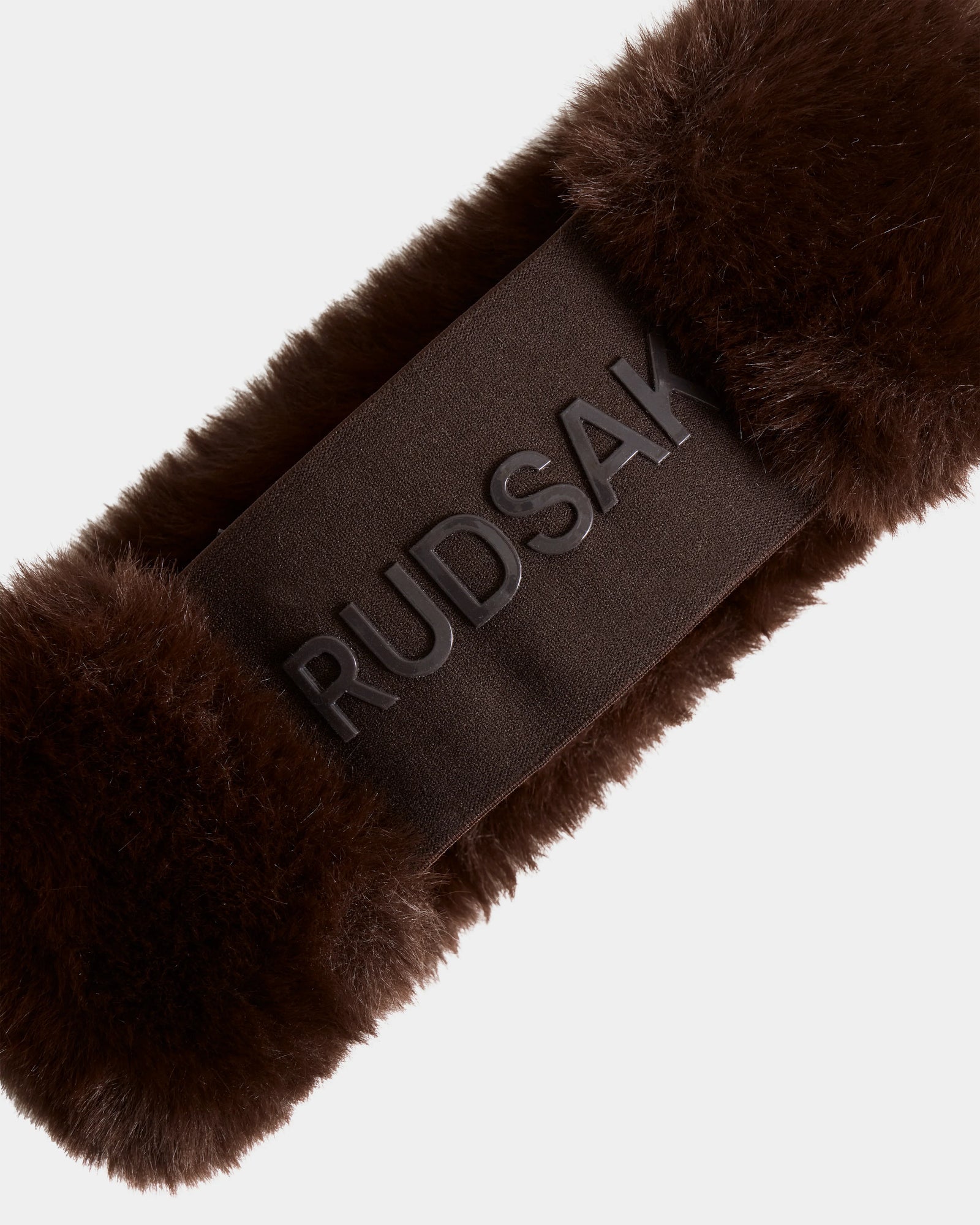 Close-up of the SANDIE - L425552 Espresso headband by Rudsak, featuring brown faux fur and a smooth dark band with raised RUDSAK text at the center. The stylish women’s winter accessory is set against a plain, light background.