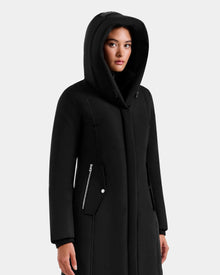 A person wears the SARINA - L125550 Black semi-fitted parka by Rudsak, featuring zippered pockets, standing against a plain light gray background with a neutral expression.