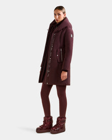 A woman stands slightly turned, wearing the Rudsak SARINA - L125550 Bordeaux long down winter coat with matching burgundy leggings and snow boots against a plain light gray background.