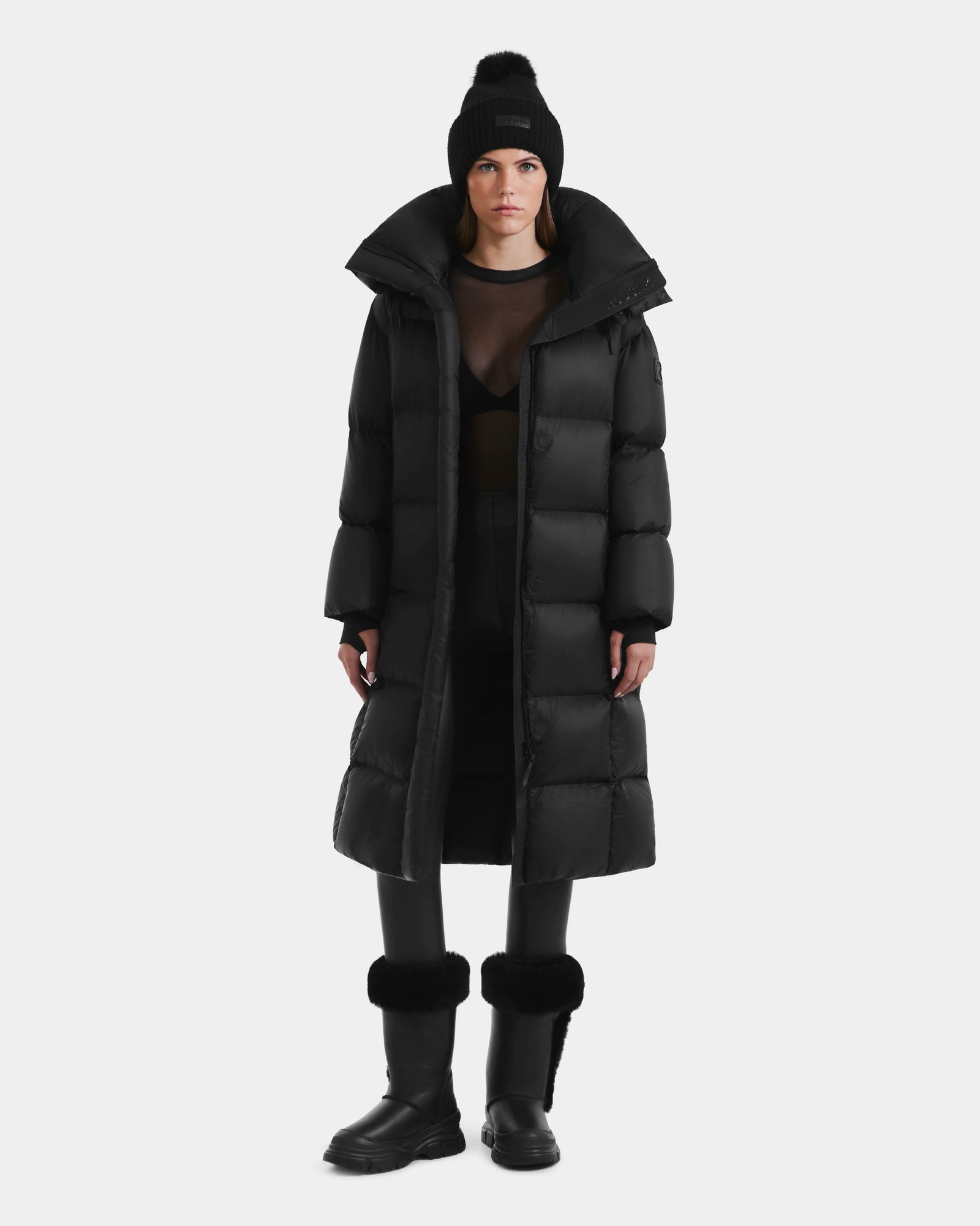 Wearing the Rudsak SASHA - 8125686 Black womens long down puffer coat with removable hood, black beanie with pom-pom, mesh top, pants, and boots, a person stands against a plain white background.