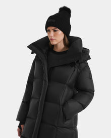 A woman wearing the Rudsak SASHA - 8125686 Black long down puffer coat and a black knit pom-pom beanie stands against a plain white background, her left hand in her coat pocket.