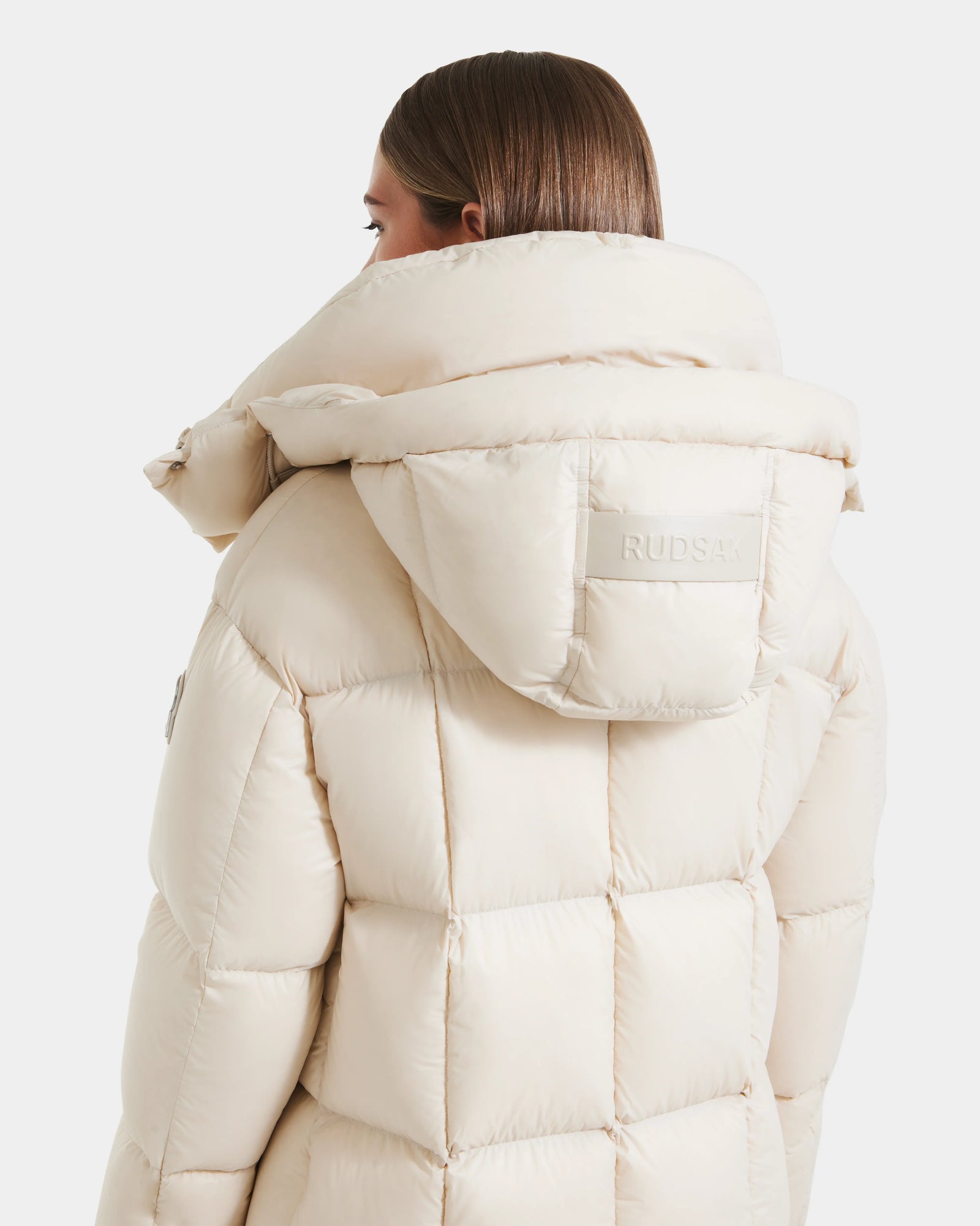 A person with straight brown hair wears the Rudsak SASHA - 8125686 Vanilla women’s long down puffer coat with a large removable hood featuring a stitched Rudsak patch on the back, set against a plain white background.