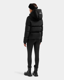 A person faces away, wearing the Rudsak STEFANIE M - L125526 Black puffer jacket with hood, black leggings, combat boots, and a black fur headband against a plain light background.