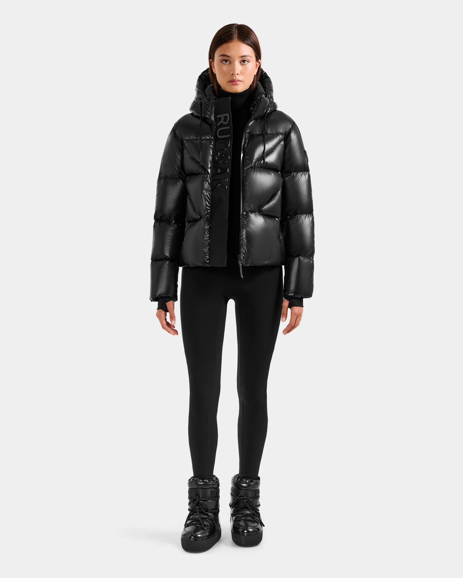 A person faces forward wearing the Rudsak STEFANIE S - L125585 Black women's shiny short puffer jacket, paired with black leggings, black boots, and a black turtleneck. The background is plain light gray.