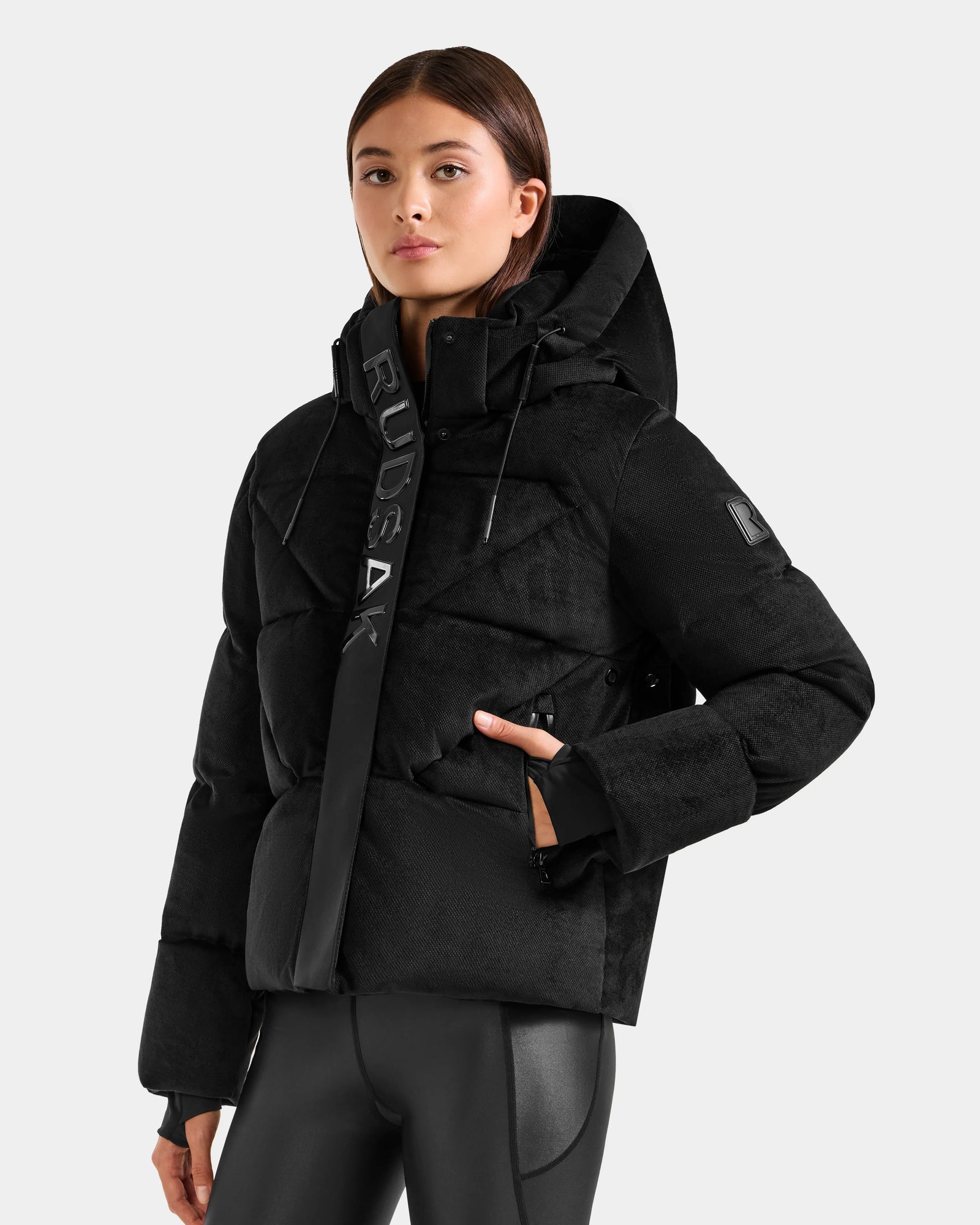 Someone wearing the Rudsak STEFANIE V - L125584 Black down puffer jacket with "RUDSAK" on the front collar stands against a light gray background. The winter coat features a high collar and zippered pockets.