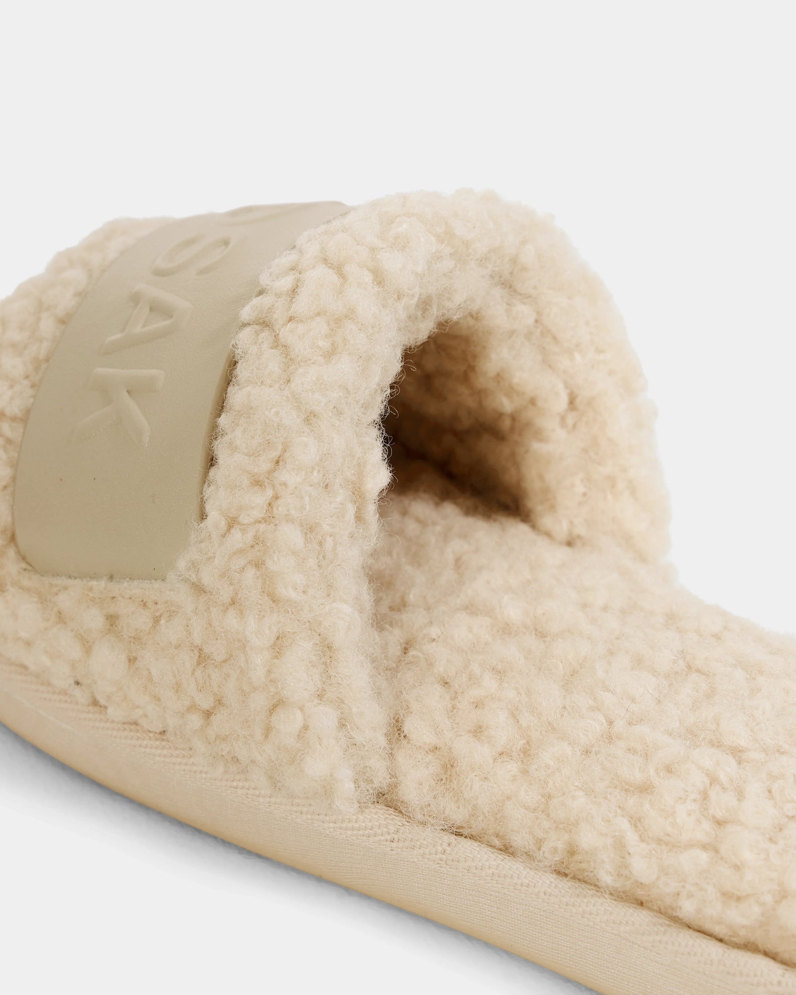 A close-up of the Rudsak TEDDY - L225517 Vanilla slipper shows its beige, wool-like faux-shearling texture and a wide, smooth vegan-leather strap. Soft and plush, it offers cozy comfort for lounging.