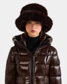 A person wears the Rudsak YURI - L425538 Espresso, a shiny dark brown puffer jacket, paired with a large faux-fur bucket hat with vegan-leather trim, looking straight at the camera against a plain light background—a chic winter accessory.