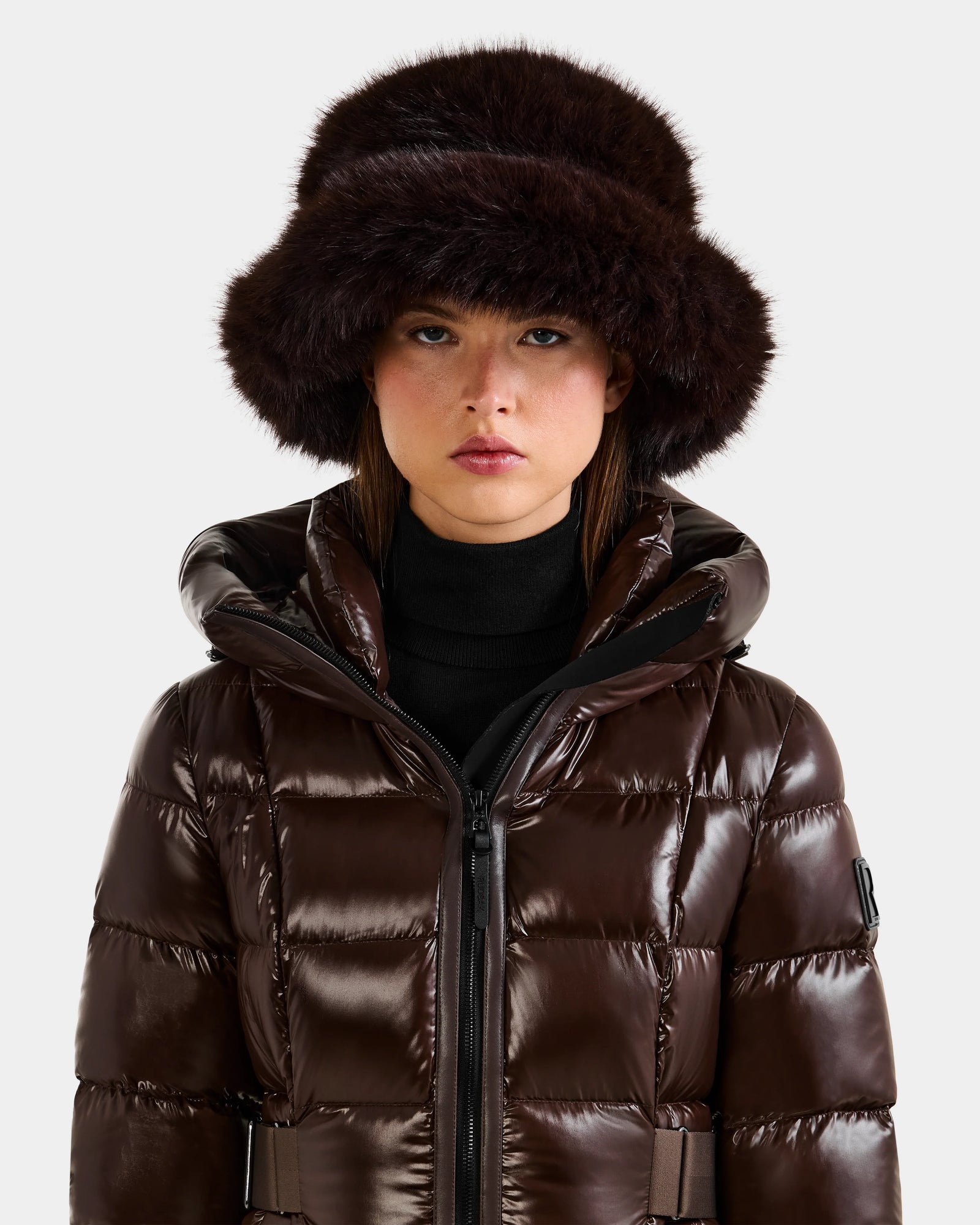 A person wears the Rudsak YURI - L425538 Espresso, a shiny dark brown puffer jacket, paired with a large faux-fur bucket hat with vegan-leather trim, looking straight at the camera against a plain light background—a chic winter accessory.